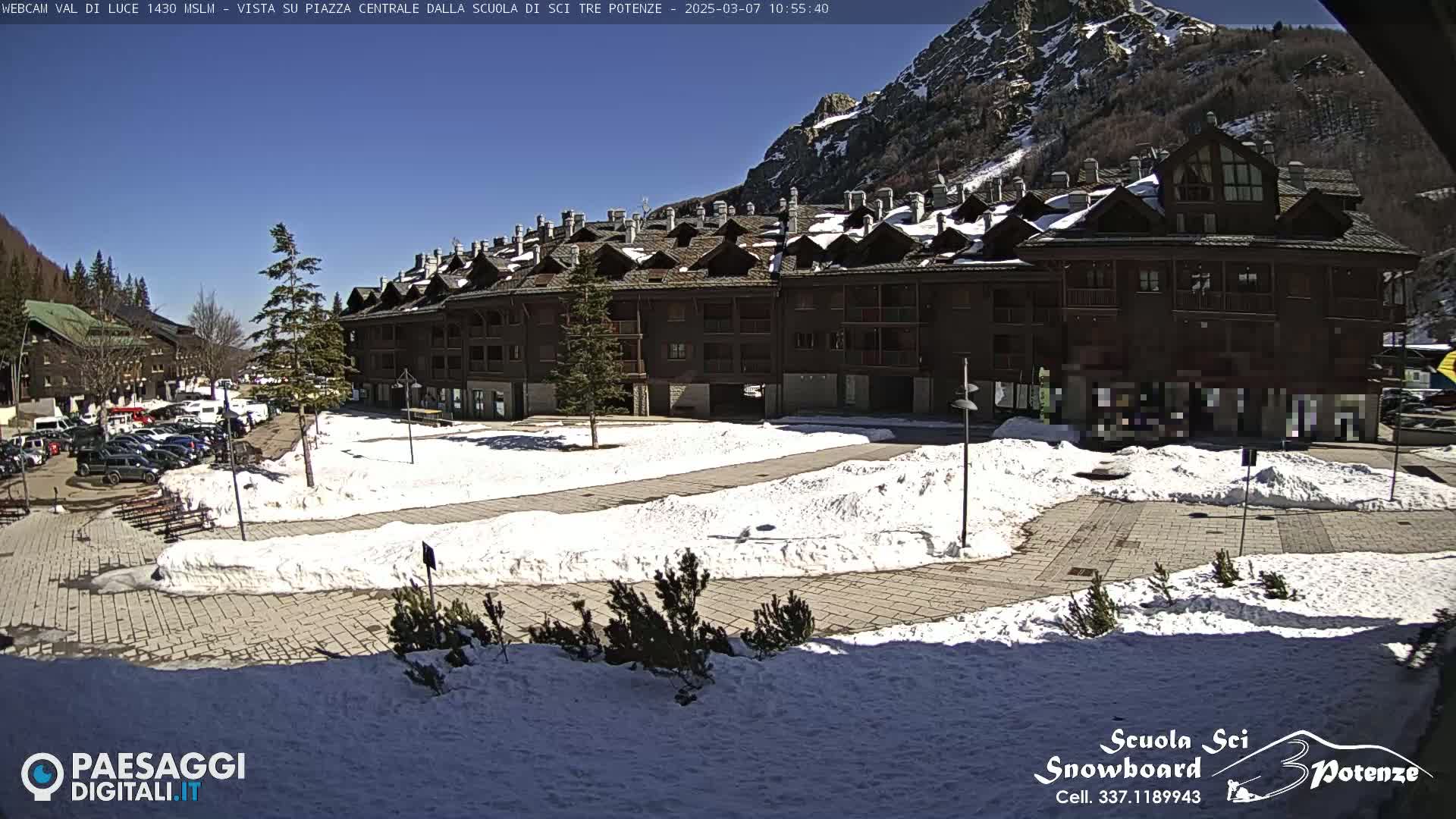 A large, multi-story building with a dark wood exterior sits in a snow-covered plaza on a sunny day in a mountainous region.