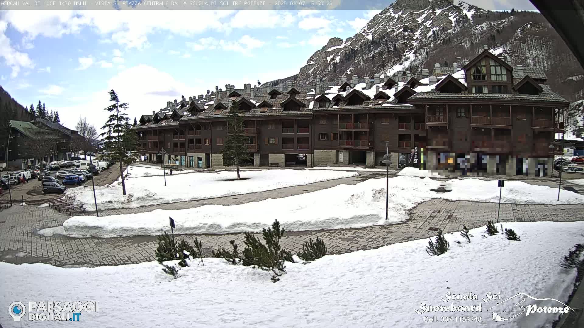 A snow-covered plaza is situated in front of a multi-story brown building, with mountains and a partly cloudy sky in the background.