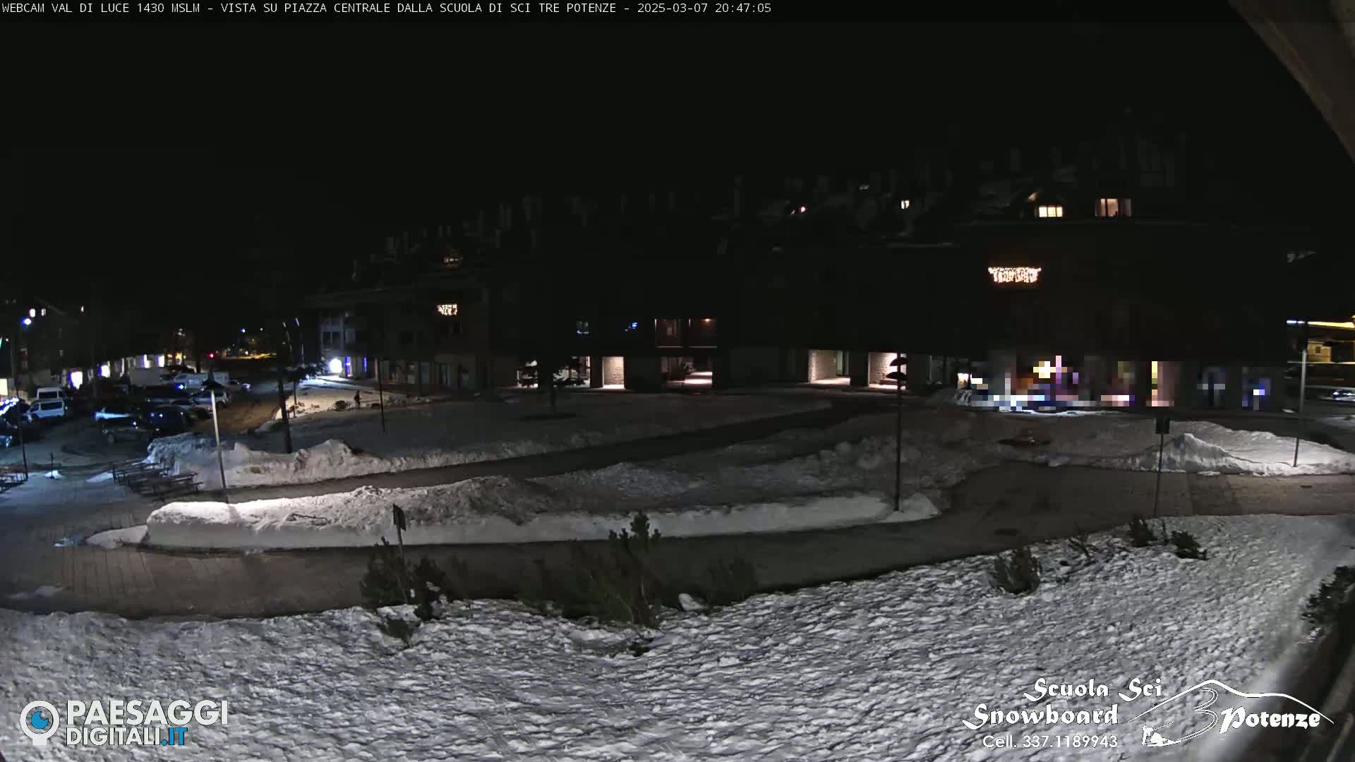 A snow-covered town square at night, illuminated by streetlights, with parked cars and buildings surrounding it.