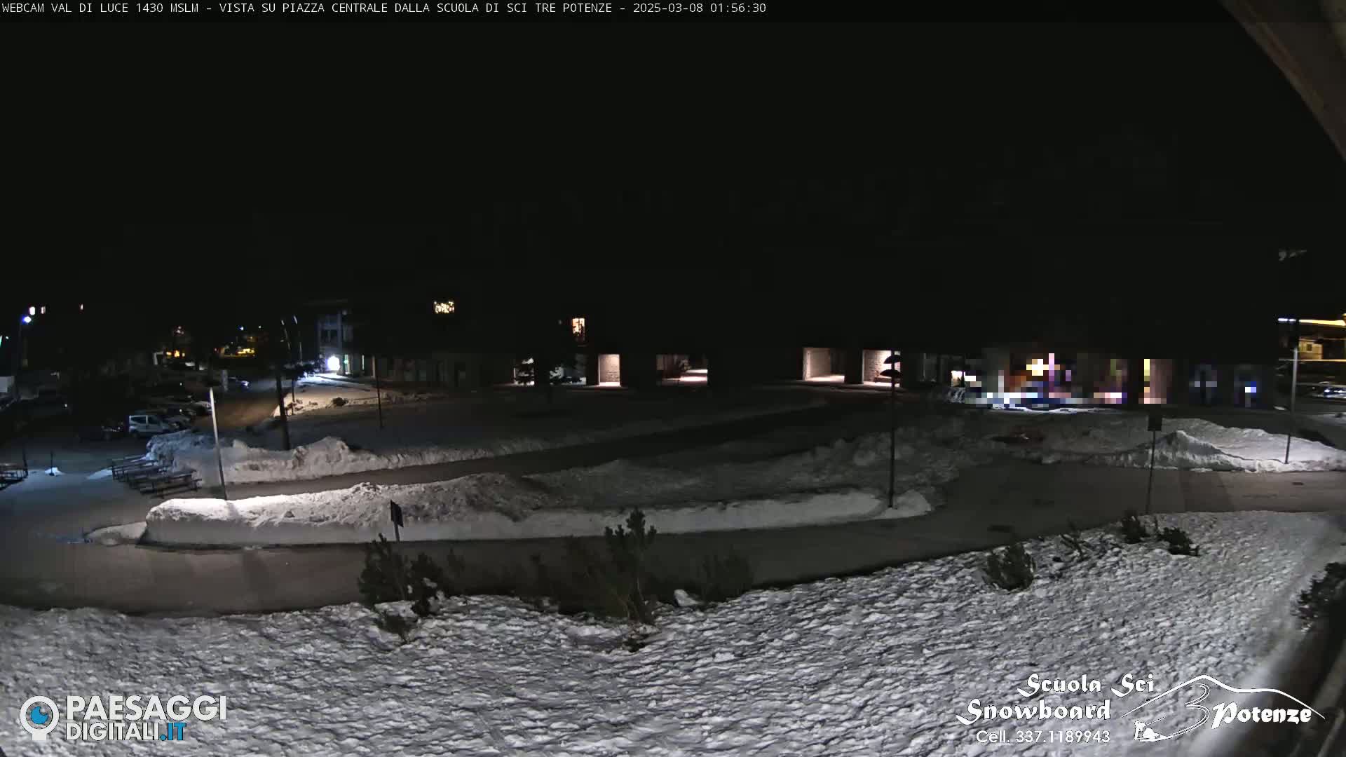 A snow-covered nighttime scene shows a paved area with buildings in the background under a dark sky.