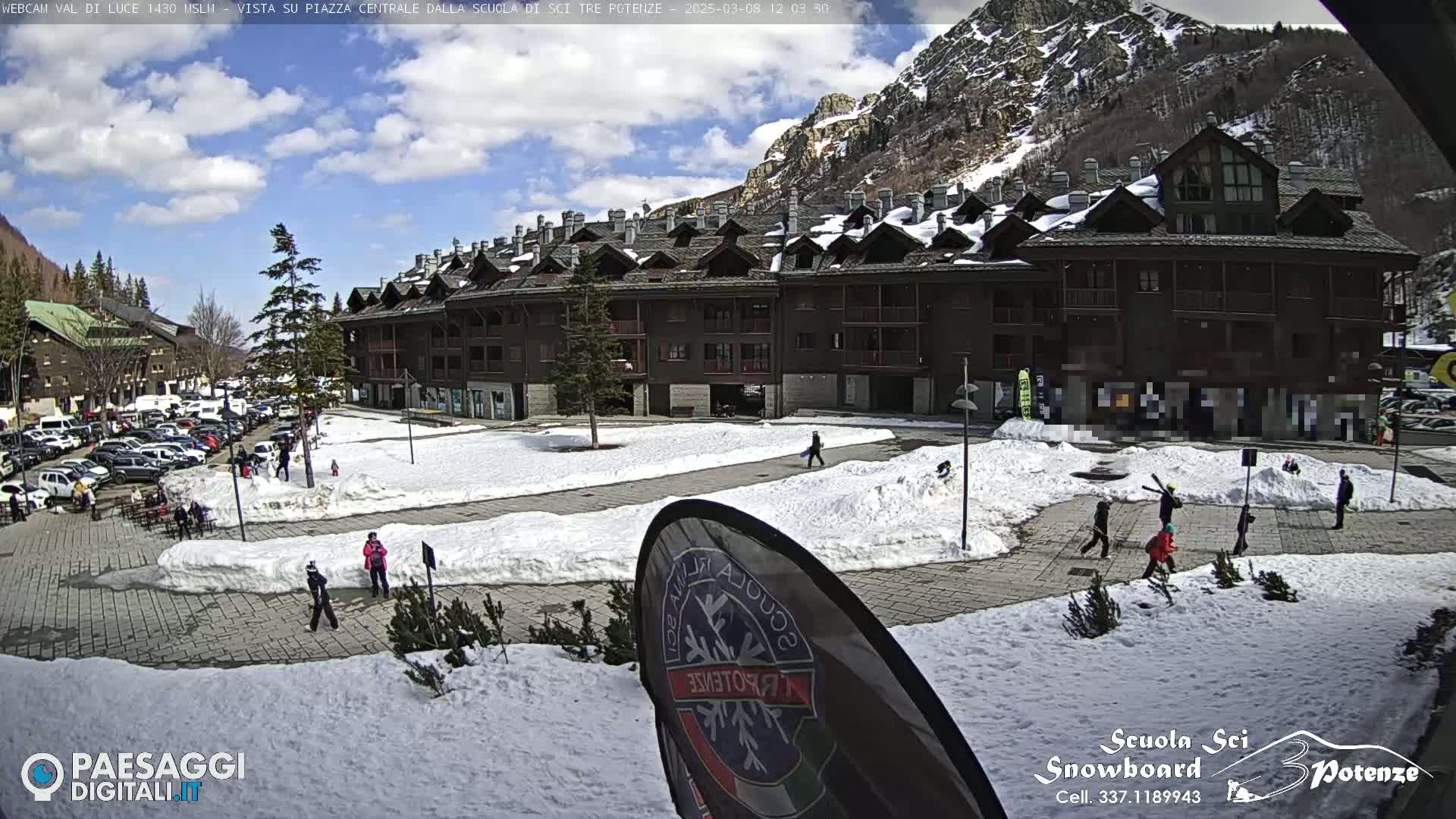 A snow-covered plaza in a mountainous region, with a multi-story building and several people walking around under a partly cloudy sky.