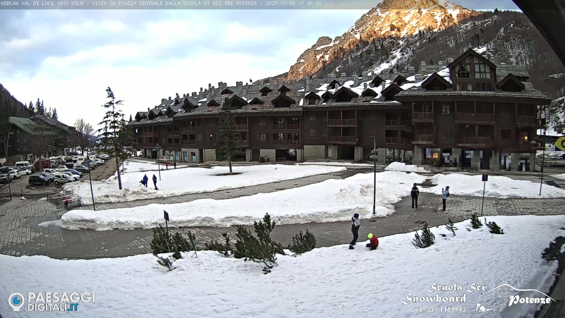 A snow-covered plaza with a multi-story building is seen in the daytime under a partly cloudy sky, with mountains in the background.
