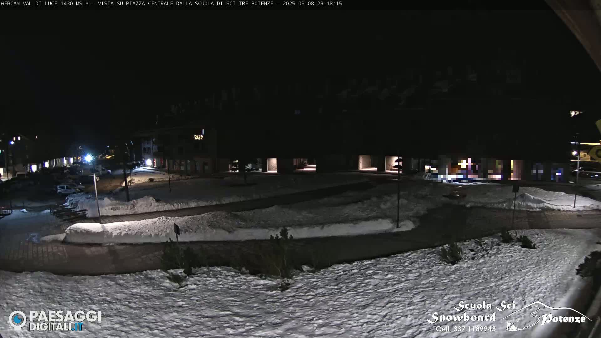 A snow-covered town square at night, illuminated by streetlights, with buildings and parked cars visible in the background.