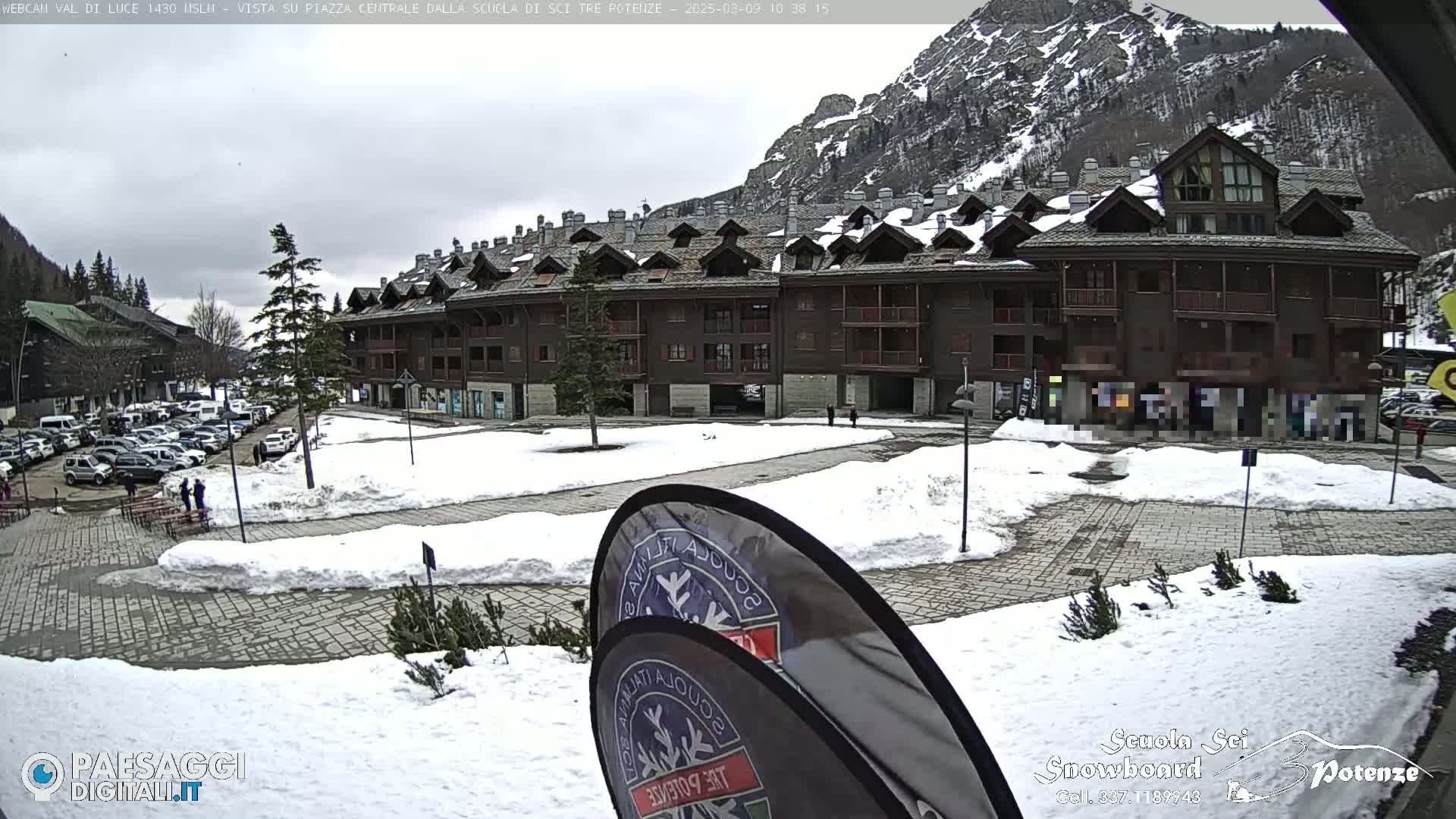 A snow-covered plaza in front of a multi-story brown building, with mountains in the background under an overcast sky.