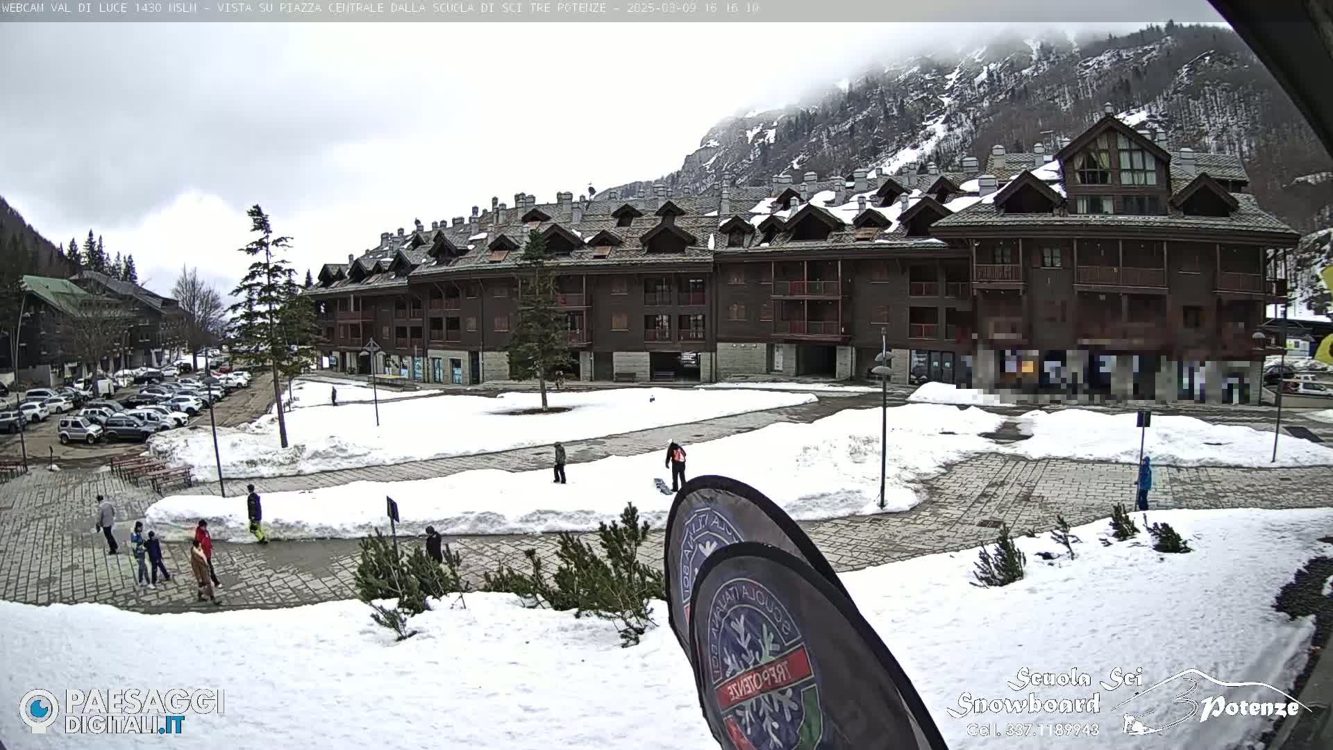 A snowy plaza with a multi-story building and several people, some of whom are snowboarding, under an overcast sky.