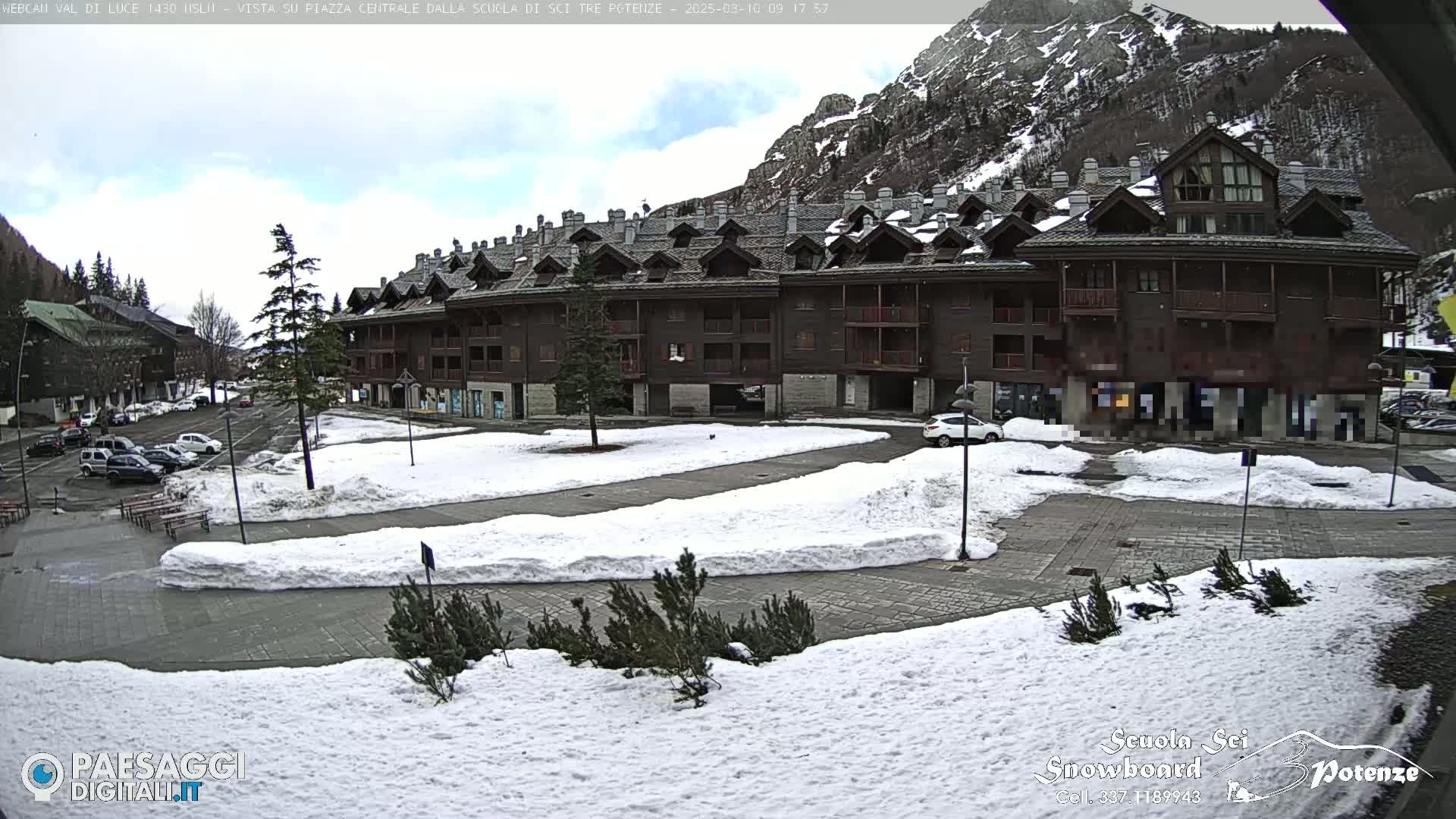 A snow-covered plaza with a large multi-story building and parked cars sits below a partly cloudy sky and mountains in the background.