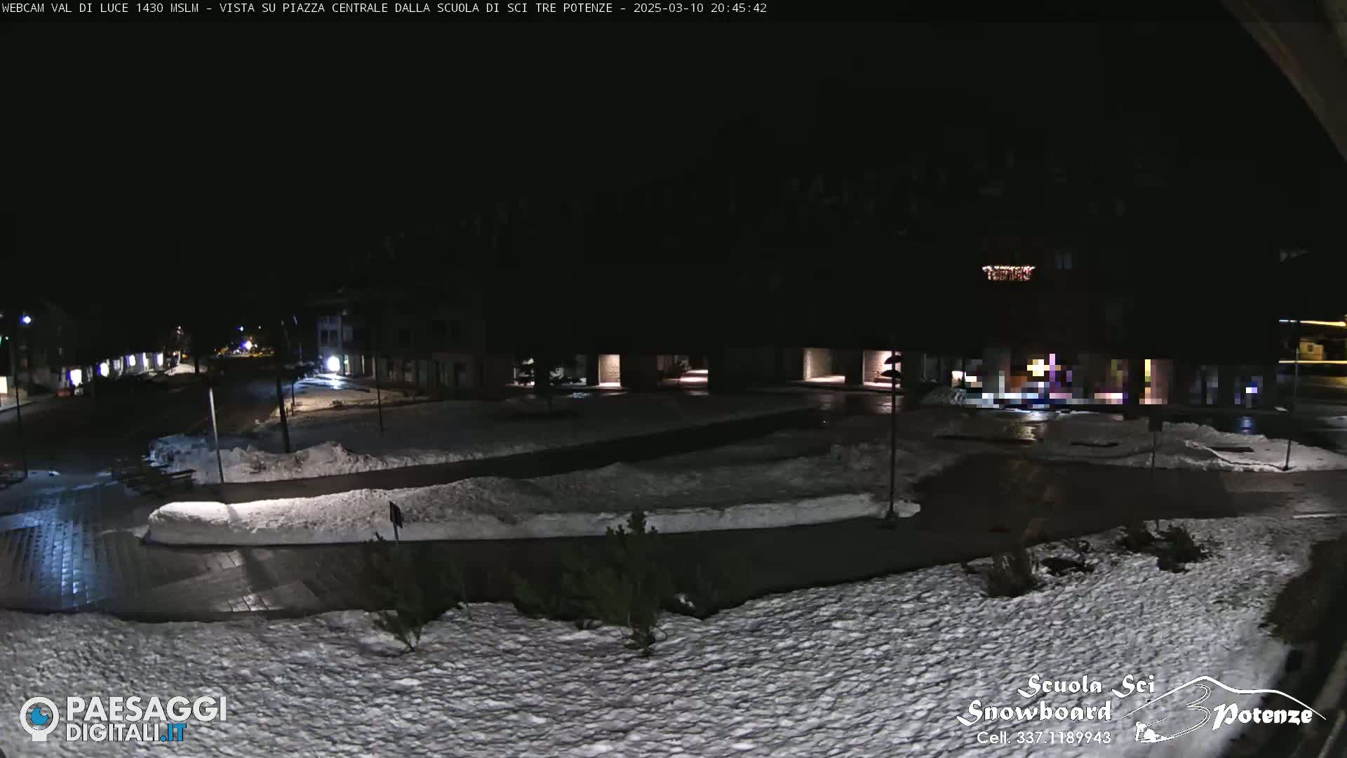A snow-covered town square at night, illuminated by streetlights, with some buildings visible in the background.