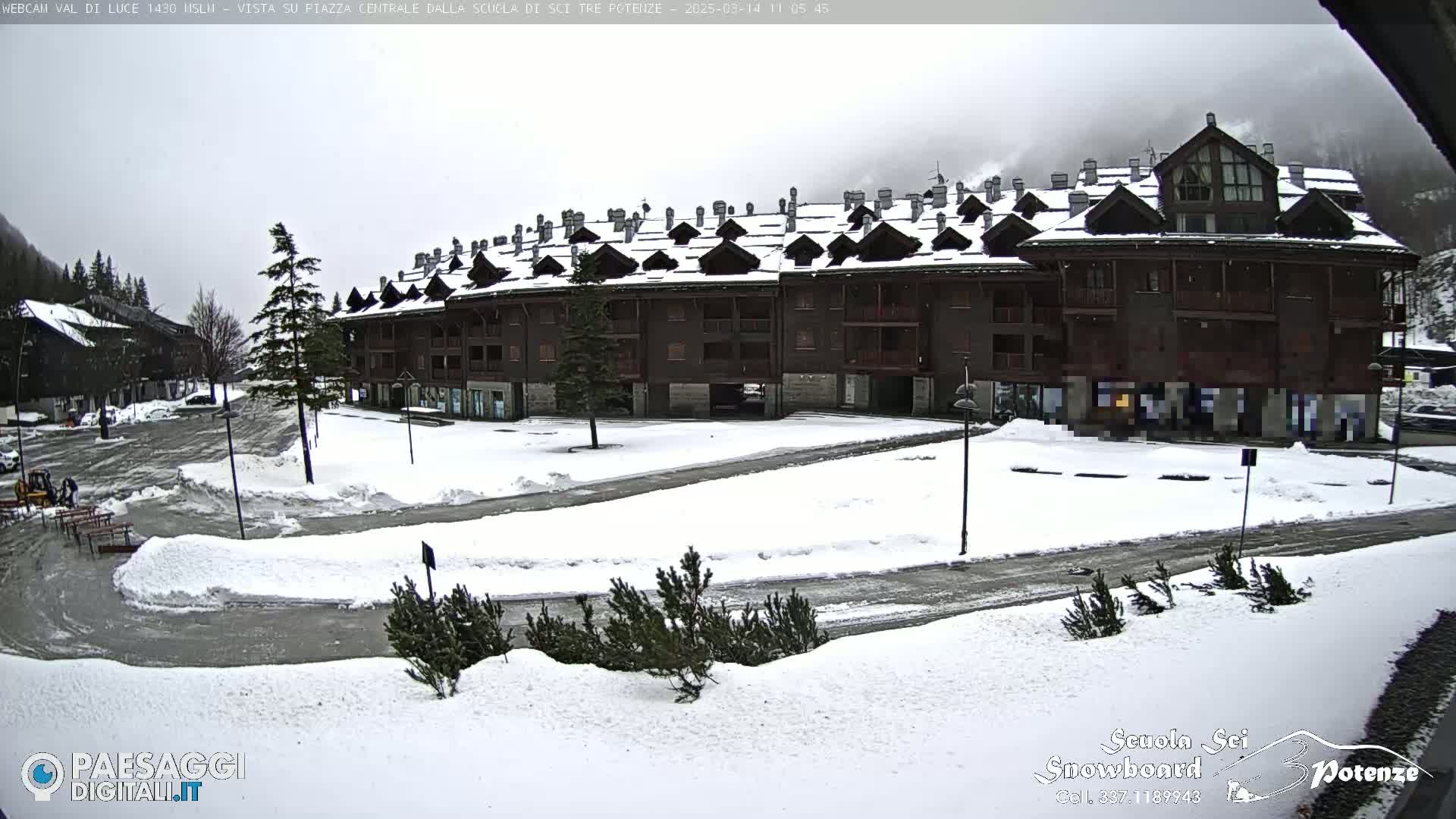 A snow-covered plaza with a multi-story brown building and some vehicles in the distance under an overcast sky.
