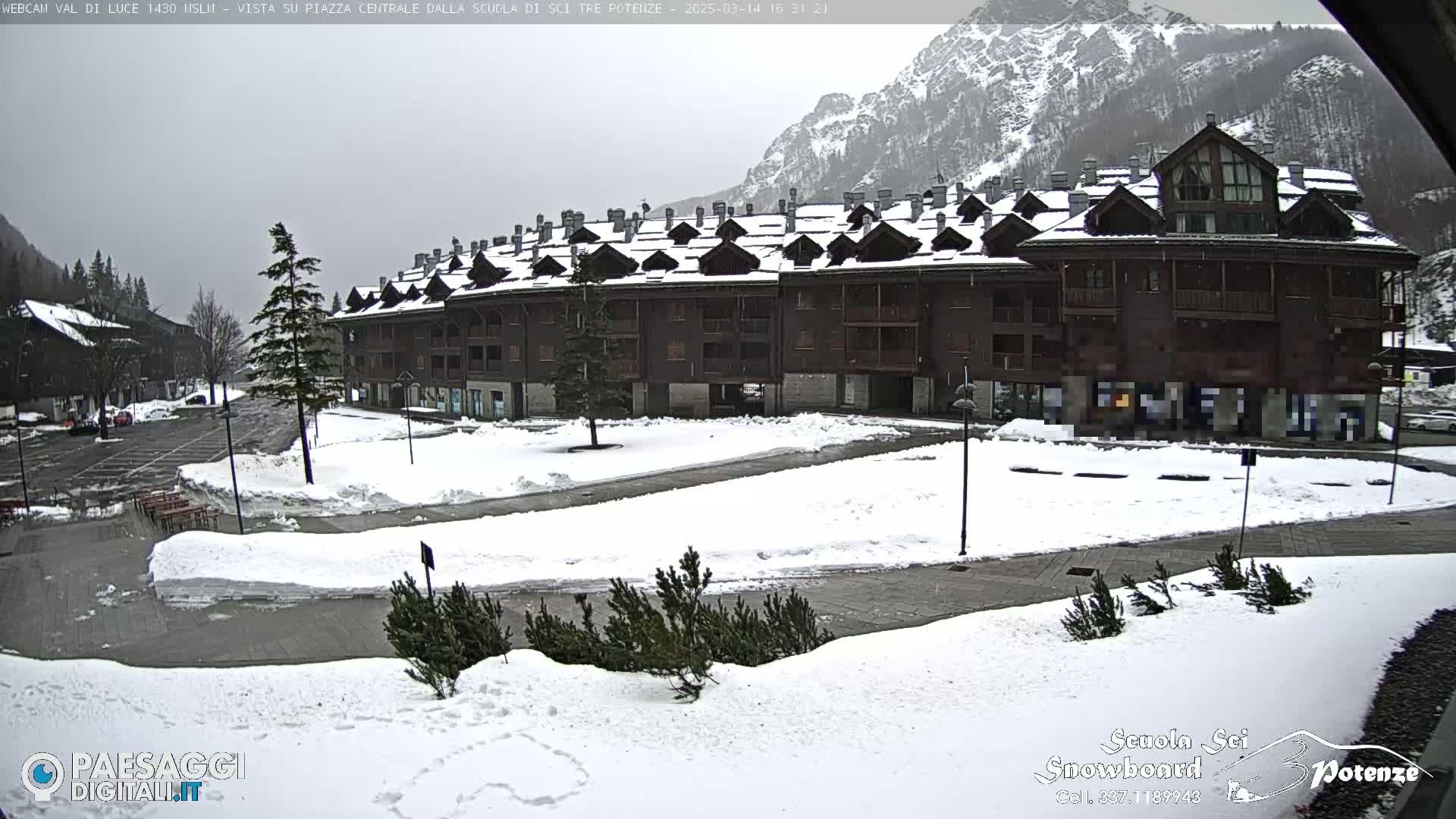 A snow-covered plaza sits in front of a multi-story brown building in a mountainous region under an overcast sky.