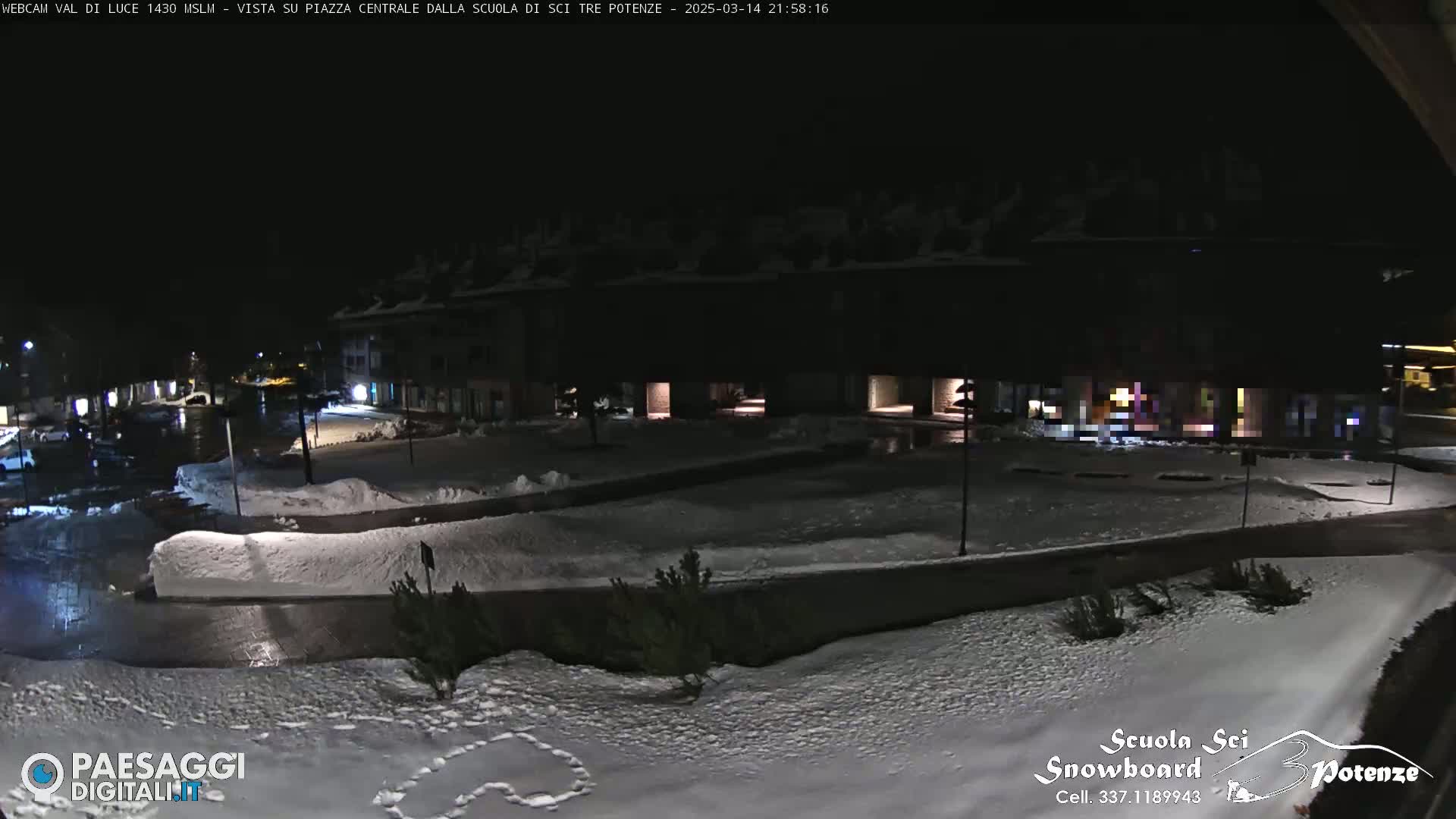 A nighttime view of a snow-covered town square, illuminated by streetlights, with buildings and some parked cars visible in the background.