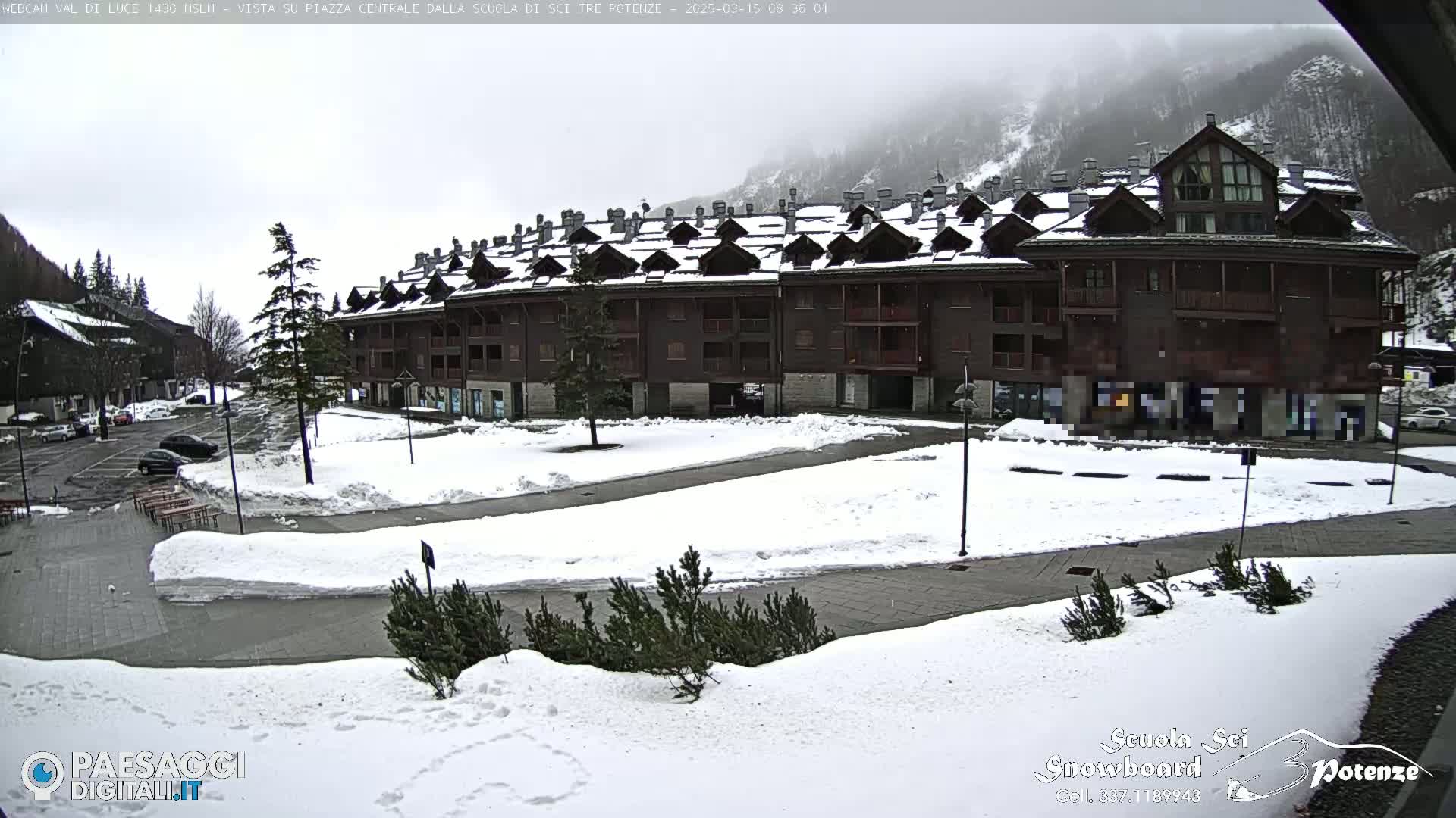 A snow-covered plaza sits in front of a large, multi-story building in a mountainous area under an overcast sky.