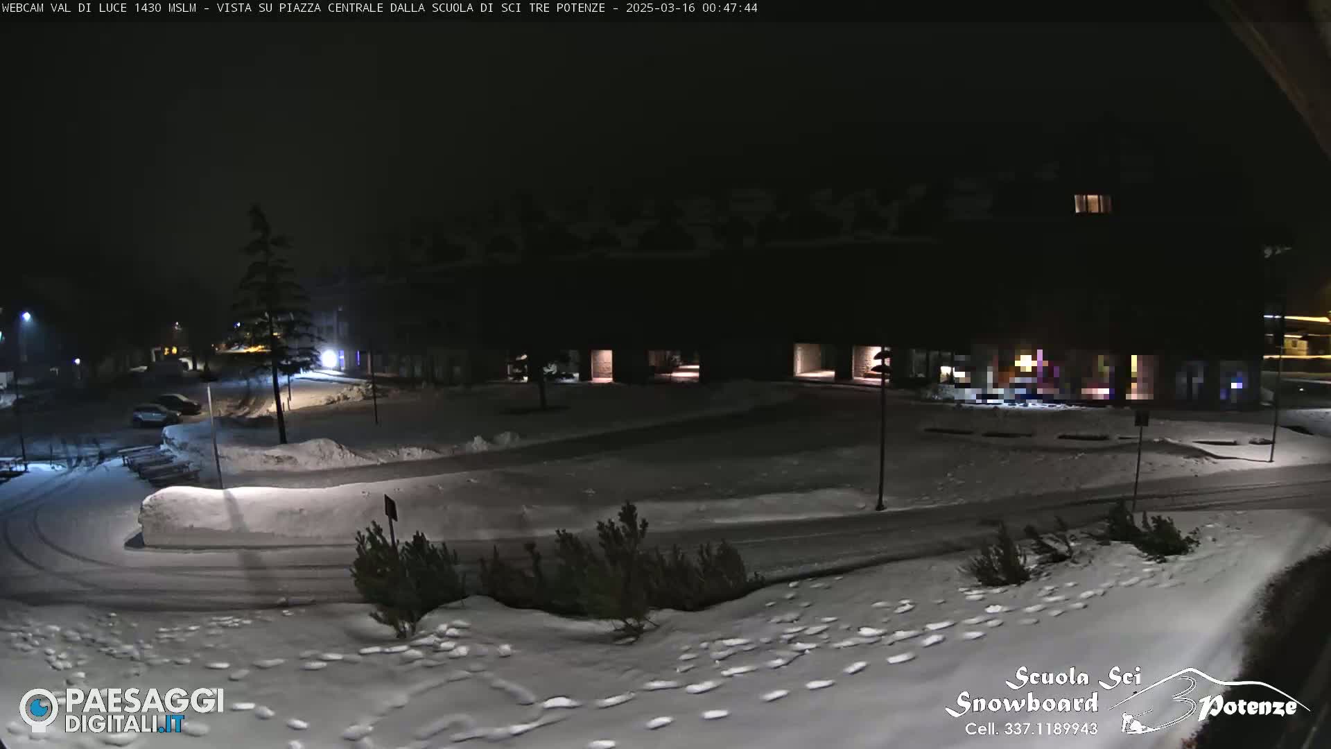 A snow-covered nighttime parking lot and plaza are illuminated by streetlights, with a few cars parked and footprints visible in the snow.