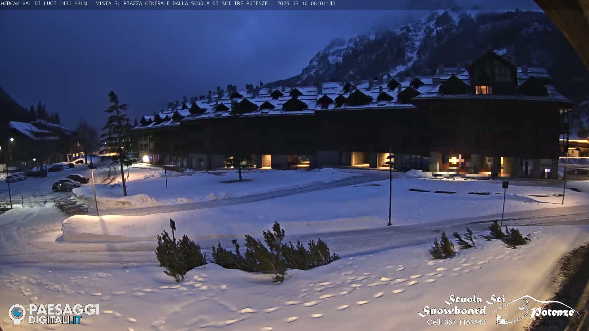 A snow-covered plaza at twilight is situated in front of a long, dark building, with a mountain visible in the background.