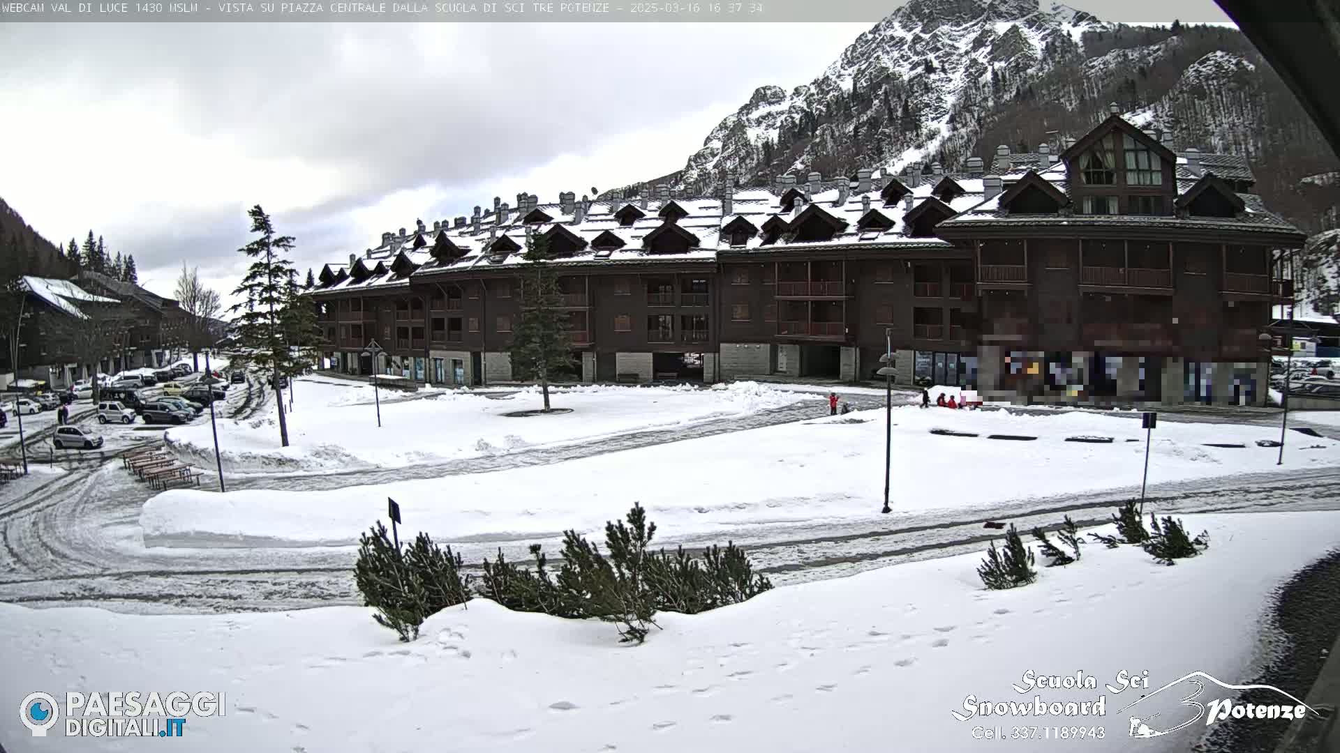 A snow-covered plaza in front of a multi-story building nestled against a mountain backdrop under a cloudy sky.