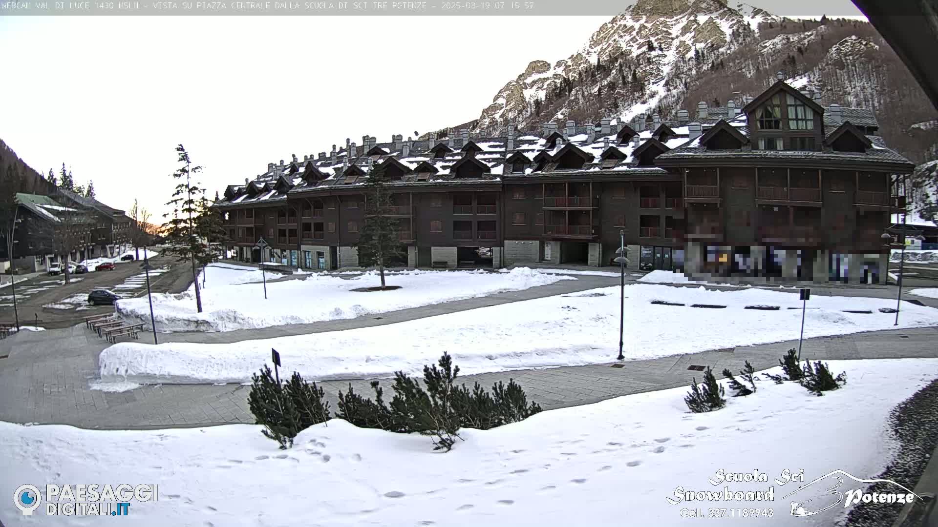 A snow-covered plaza sits in front of a multi-story building nestled in a mountain valley under a clear, bright sky.