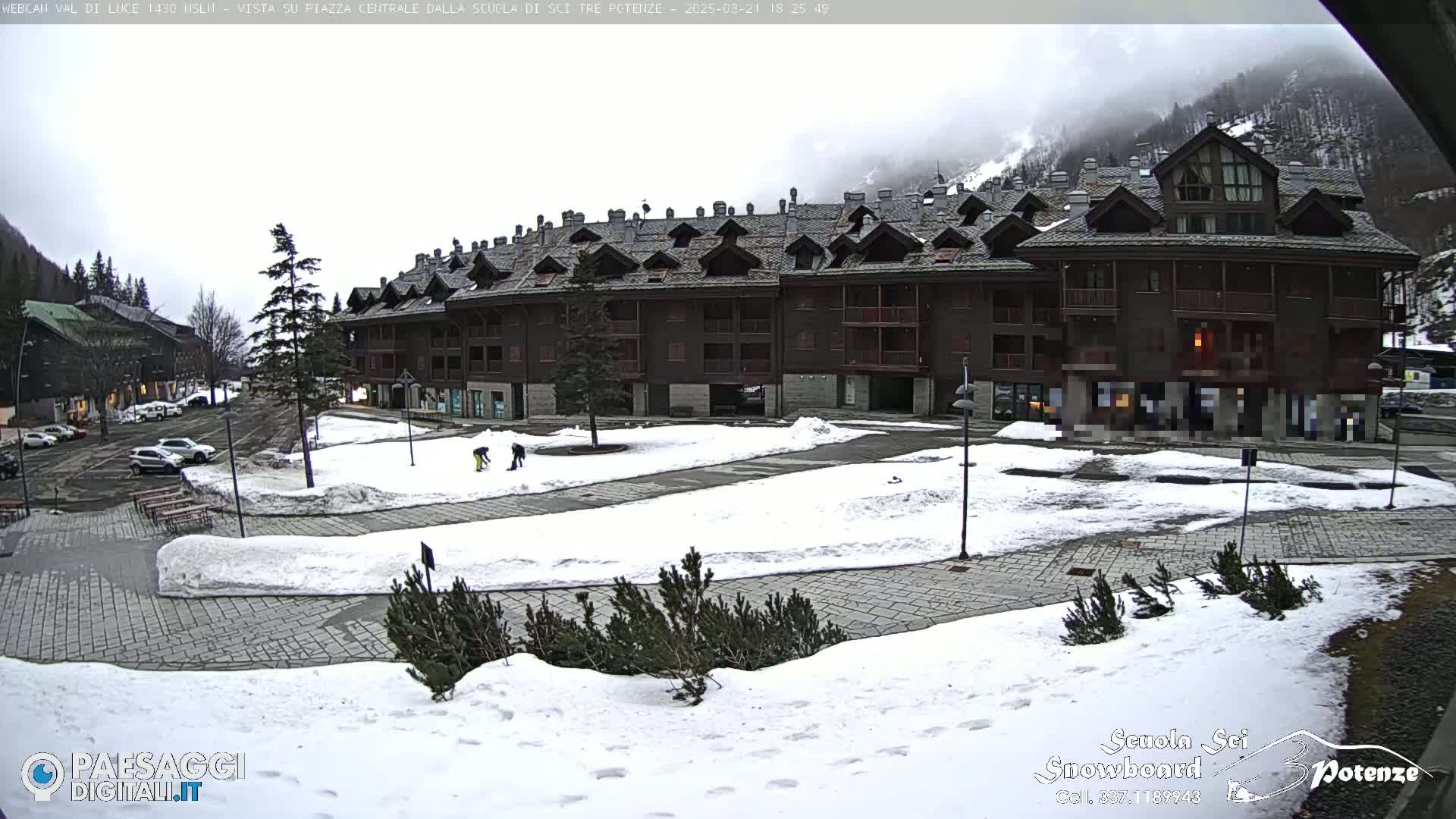 A snow-covered plaza sits in front of a large, multi-story wood-and-stone building, with overcast skies and mountains in the background.