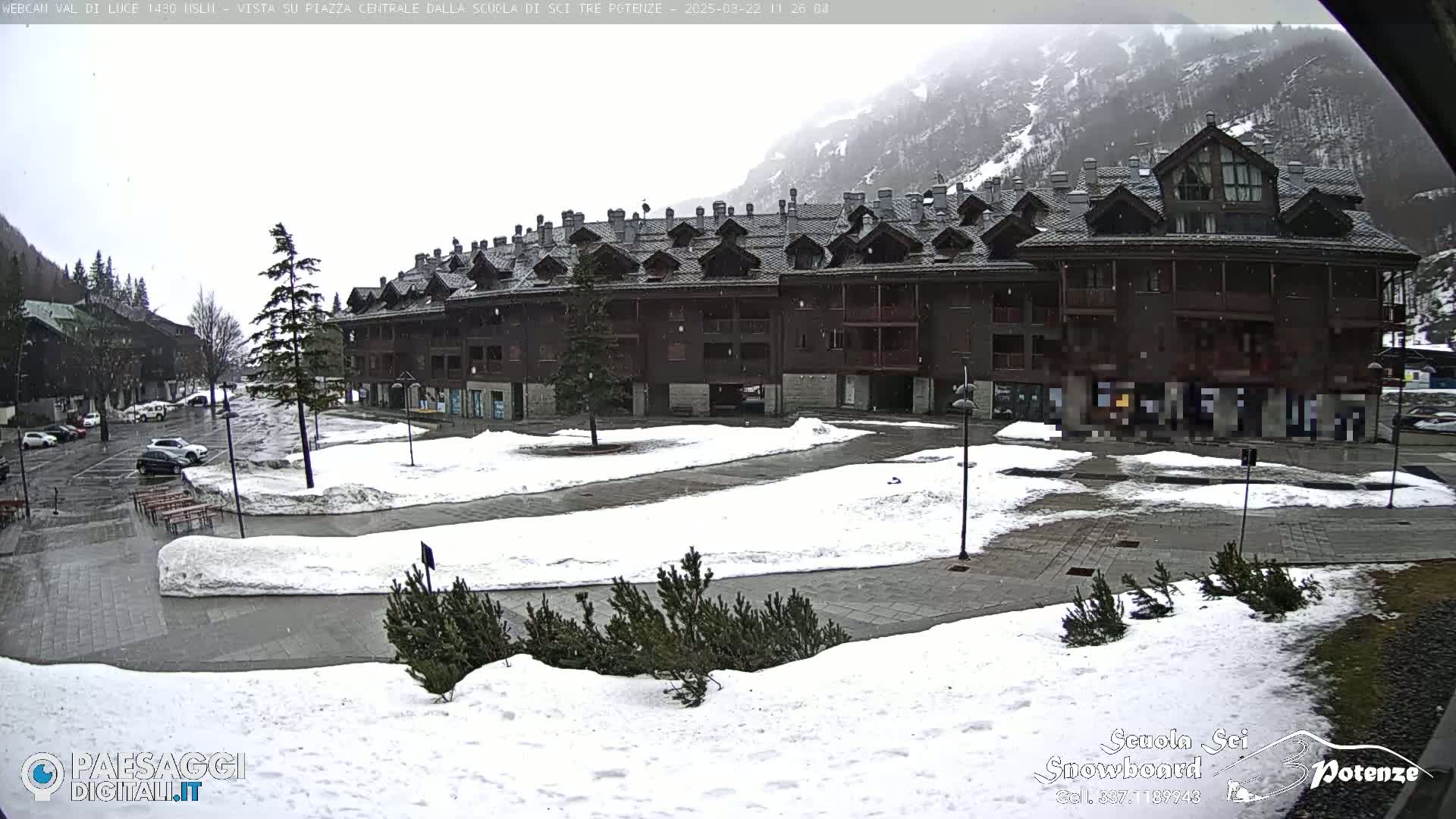 A large, dark-brown multi-story building sits in a snowy square with some cars parked nearby under an overcast sky.