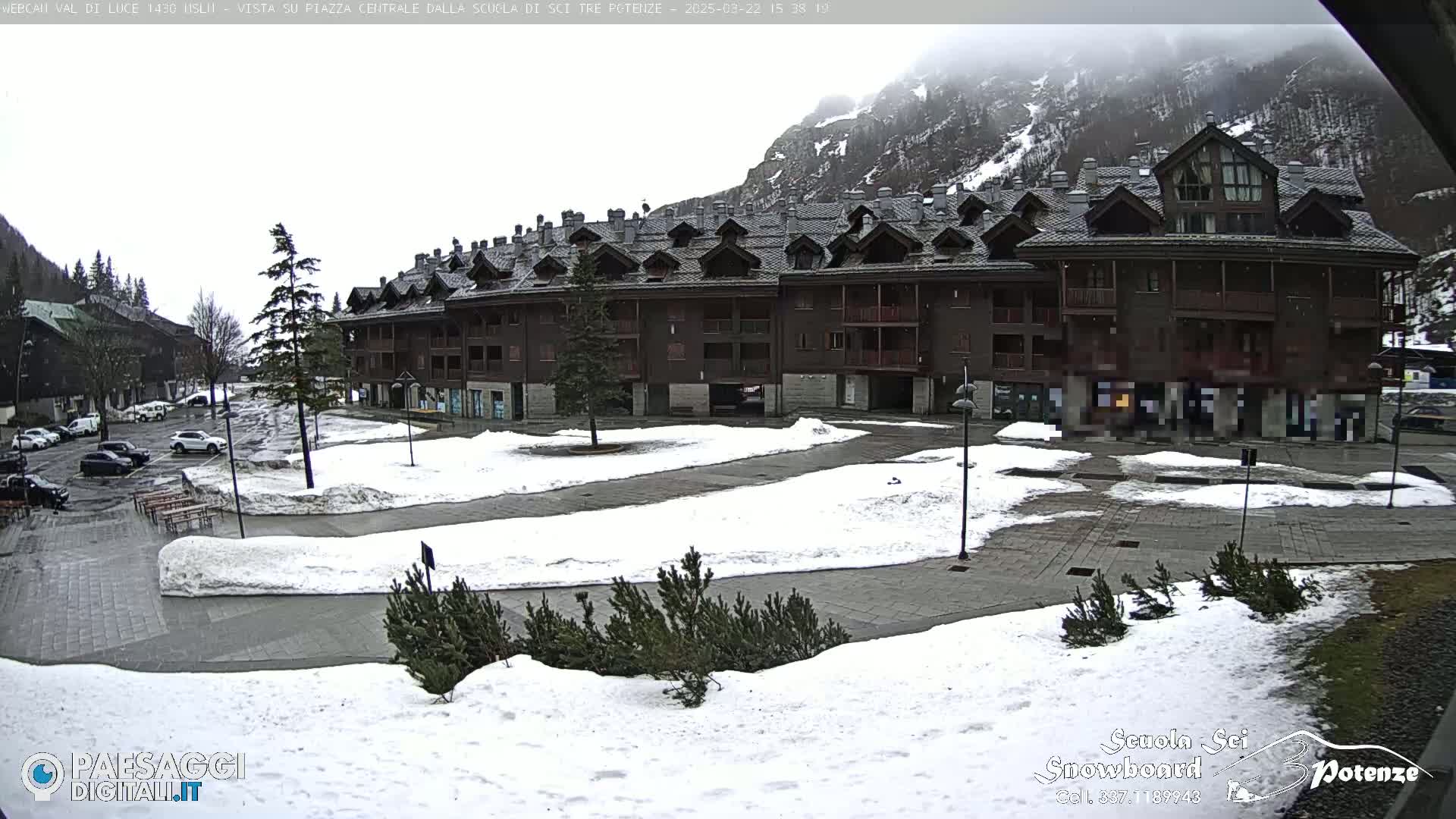 A snow-covered plaza with a large brown building sits in front of a mountain under an overcast sky.