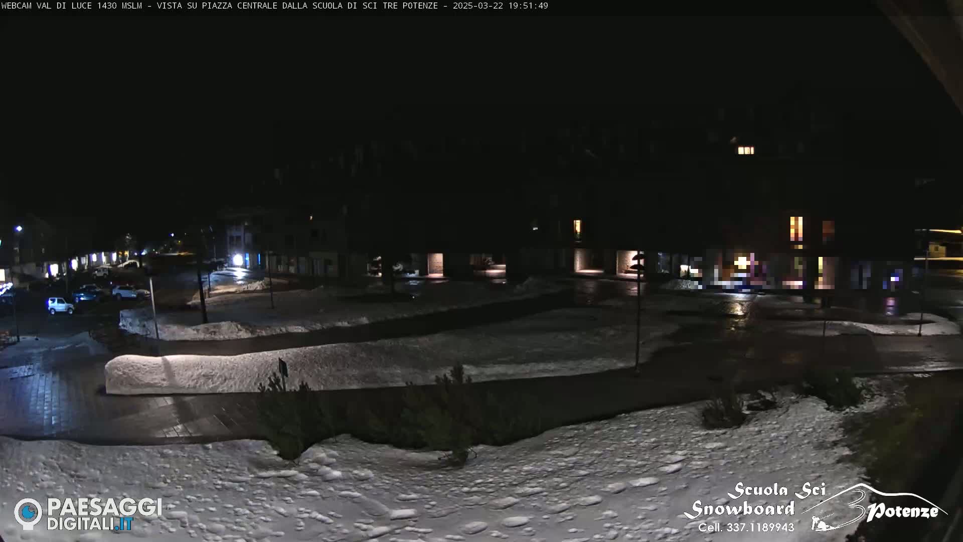 A nighttime view of a snow-covered town square with parked cars and buildings dimly lit.