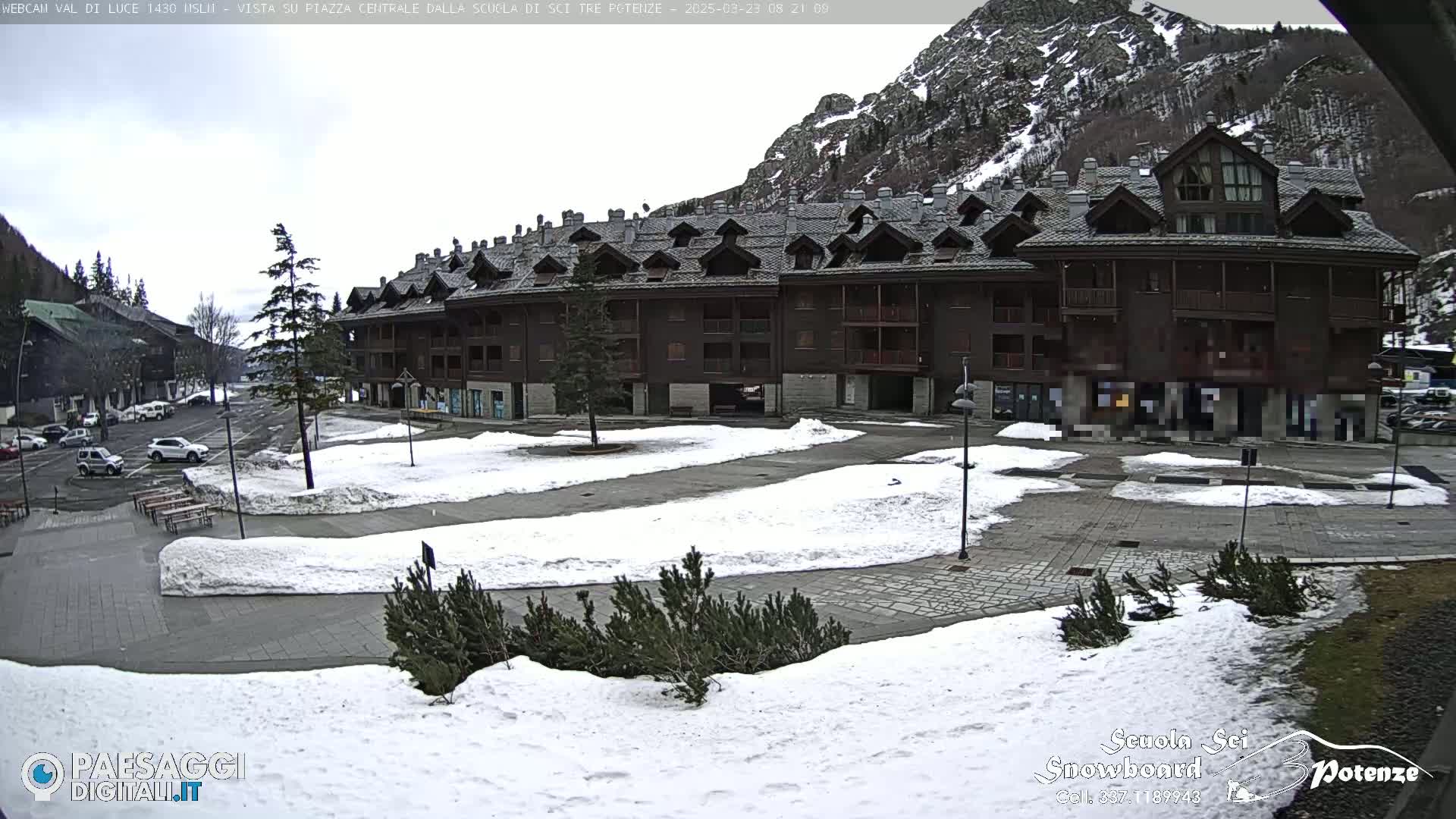 A snow-covered town square is situated in front of a large, dark-brown multi-story building, with mountains in the background under an overcast sky.