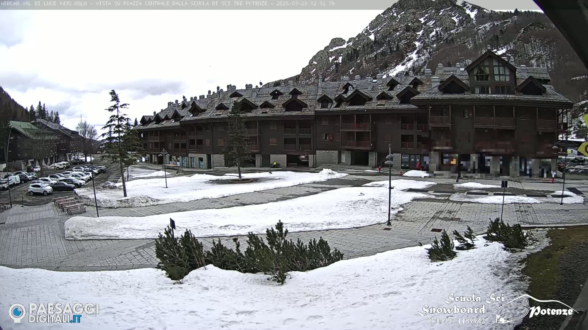 A snow-covered plaza sits in front of a multi-story brown building, with mountains and a parking lot visible in the background under a cloudy sky.