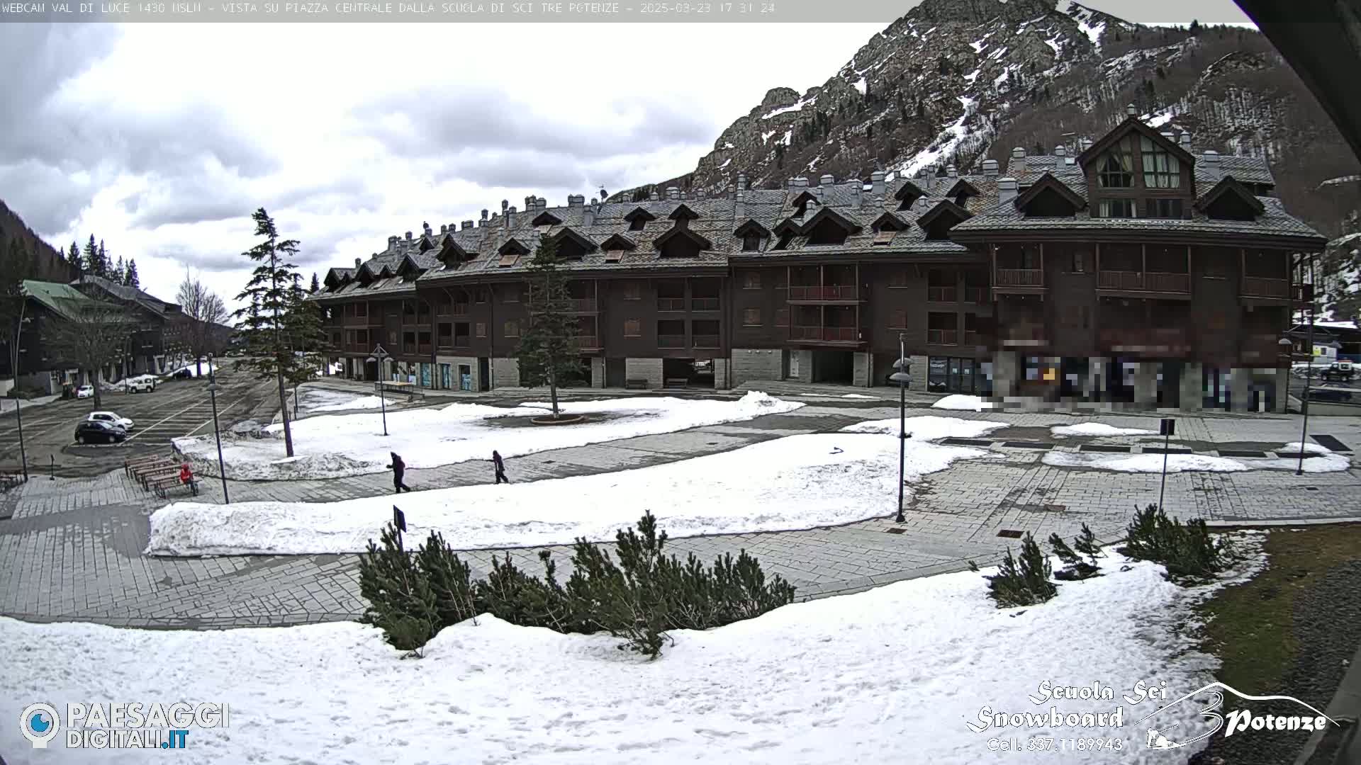 A multi-story brown building sits in a snow-dusted plaza, with a mountain backdrop under a cloudy sky.