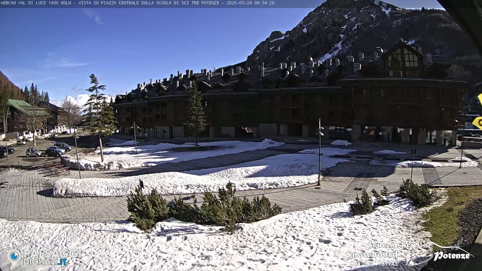 A sunny day in a snow-covered mountain town square, featuring a large multi-story building and several parked cars.