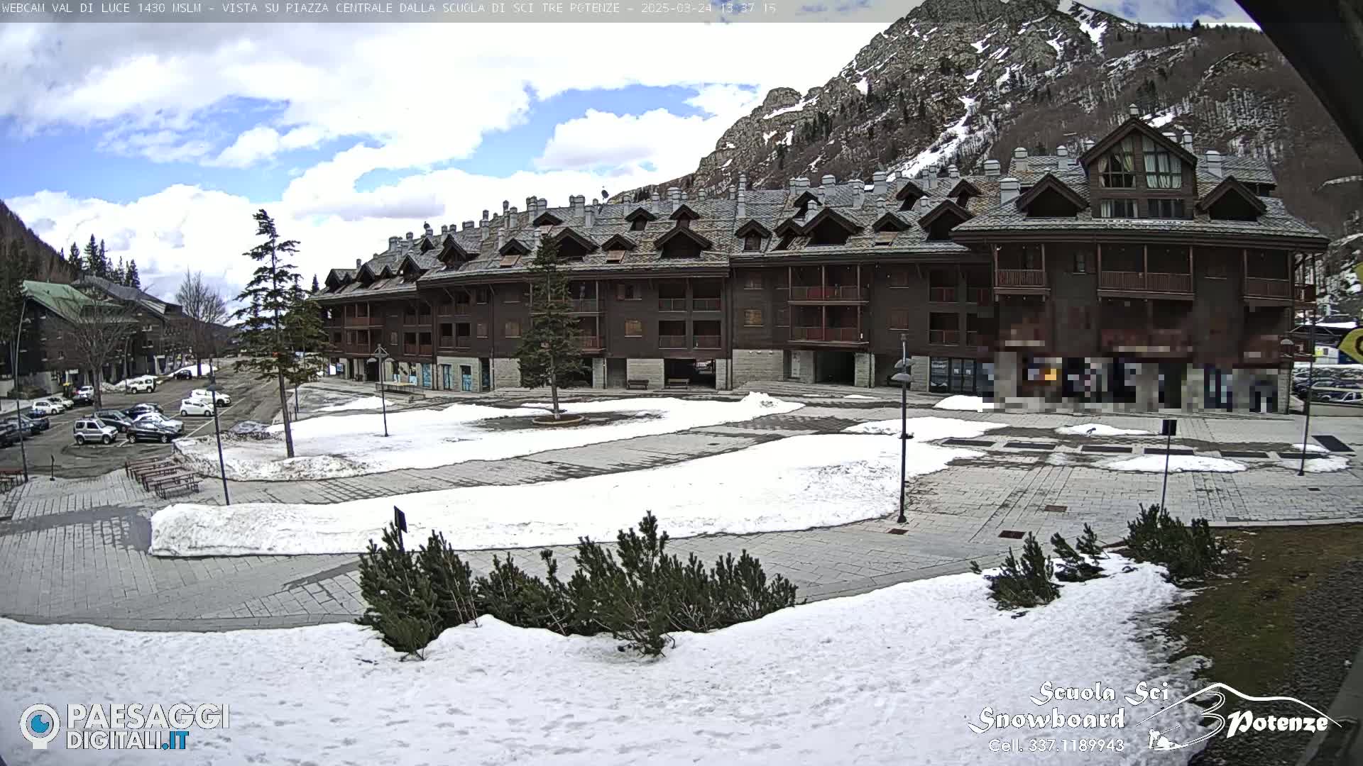 A mostly sunny day with patchy snow covers a paved plaza in front of a multi-story brown building nestled in a mountain valley.