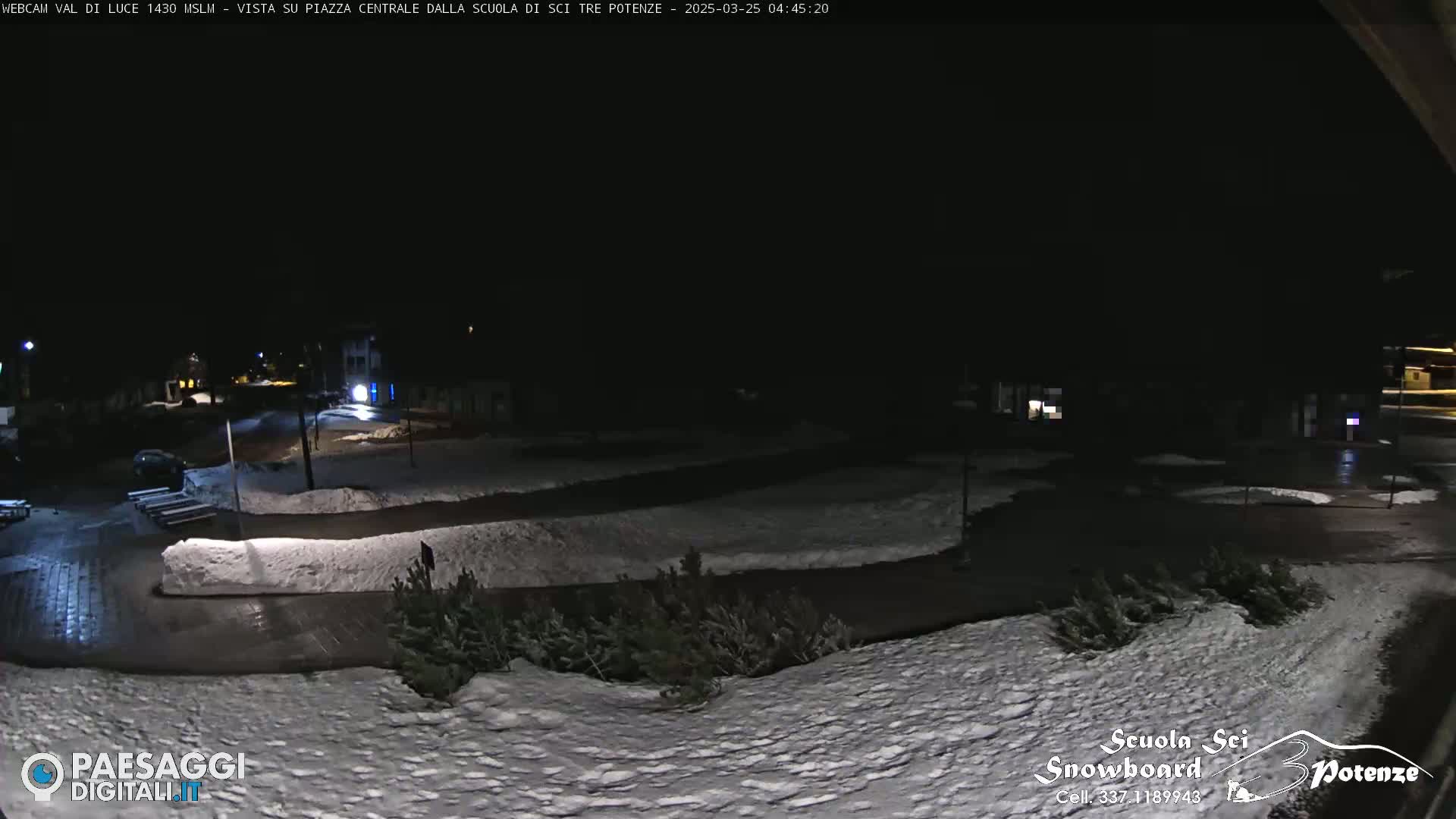 A snow-covered nighttime scene shows a paved area with some parked cars, low shrubs, and buildings in the distance under dark skies.