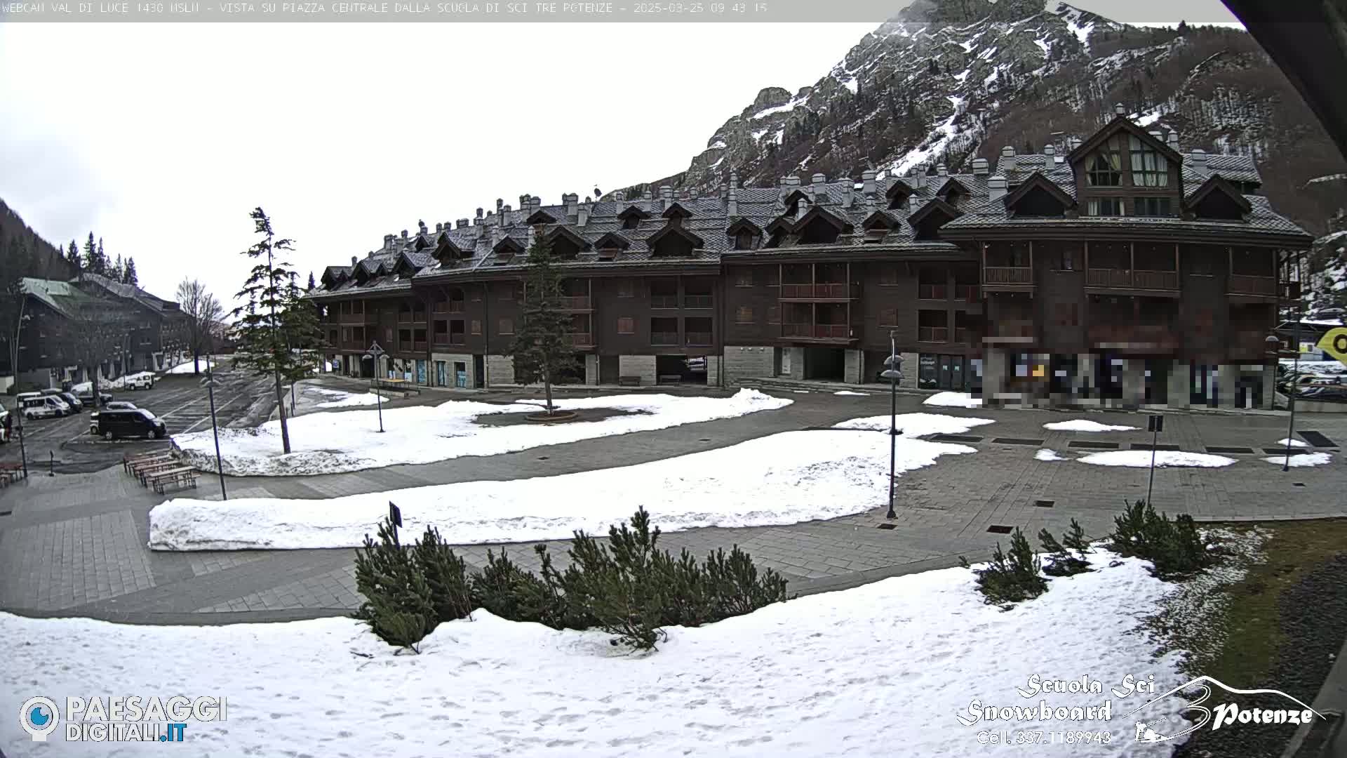 A snow-dusted plaza in front of a large, multi-story building, nestled at the base of a mountain under an overcast sky.