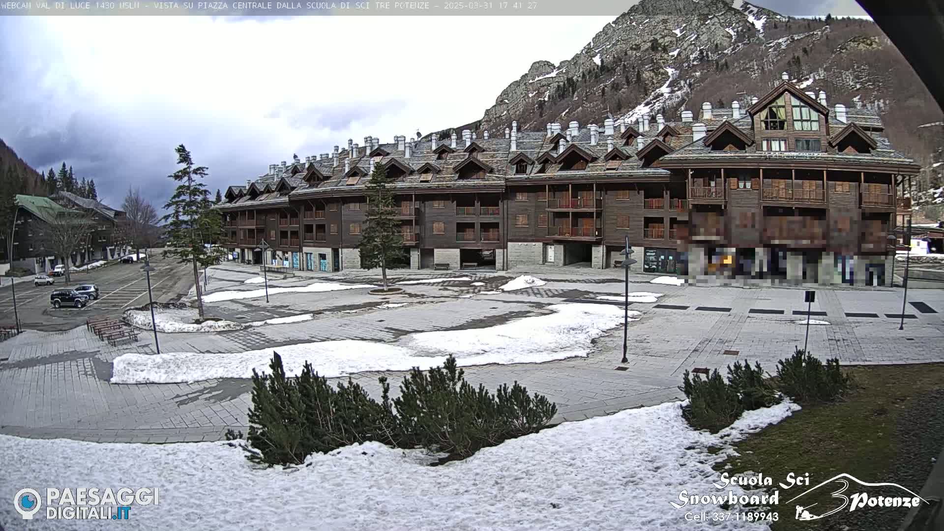 A large, multi-story, wood-sided building overlooks a snow-dusted plaza in a mountainous area under a cloudy sky.