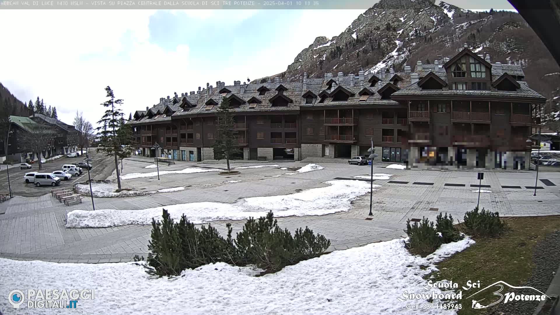 A paved square with patches of snow, surrounded by a multi-story brown building and mountains under a partly cloudy sky.