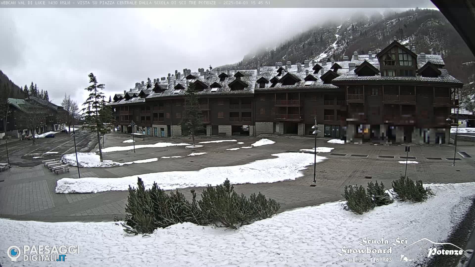 A mostly empty plaza with patches of snow, surrounded by a multi-story building and mountains under an overcast sky.