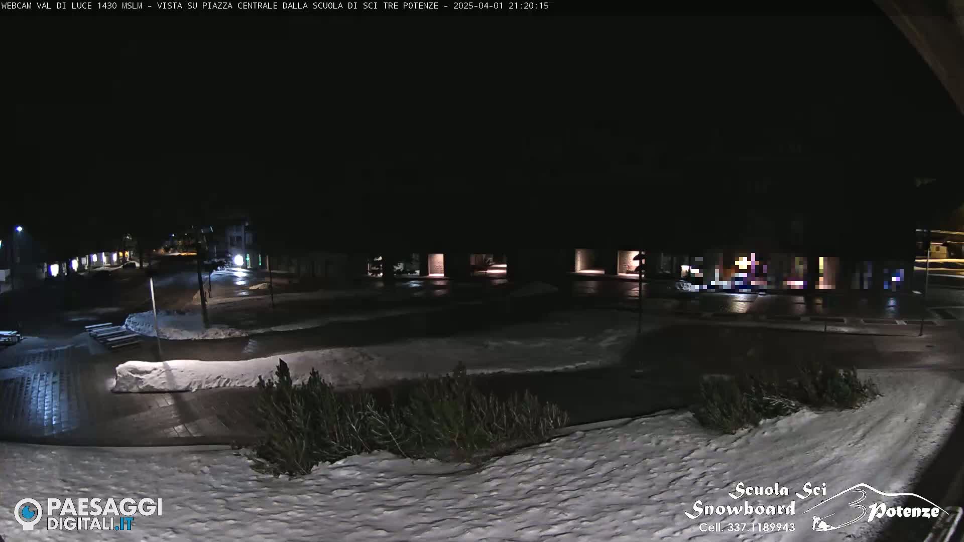 A nighttime view of a snow-covered town square with dimly lit buildings and a road, under a dark sky.