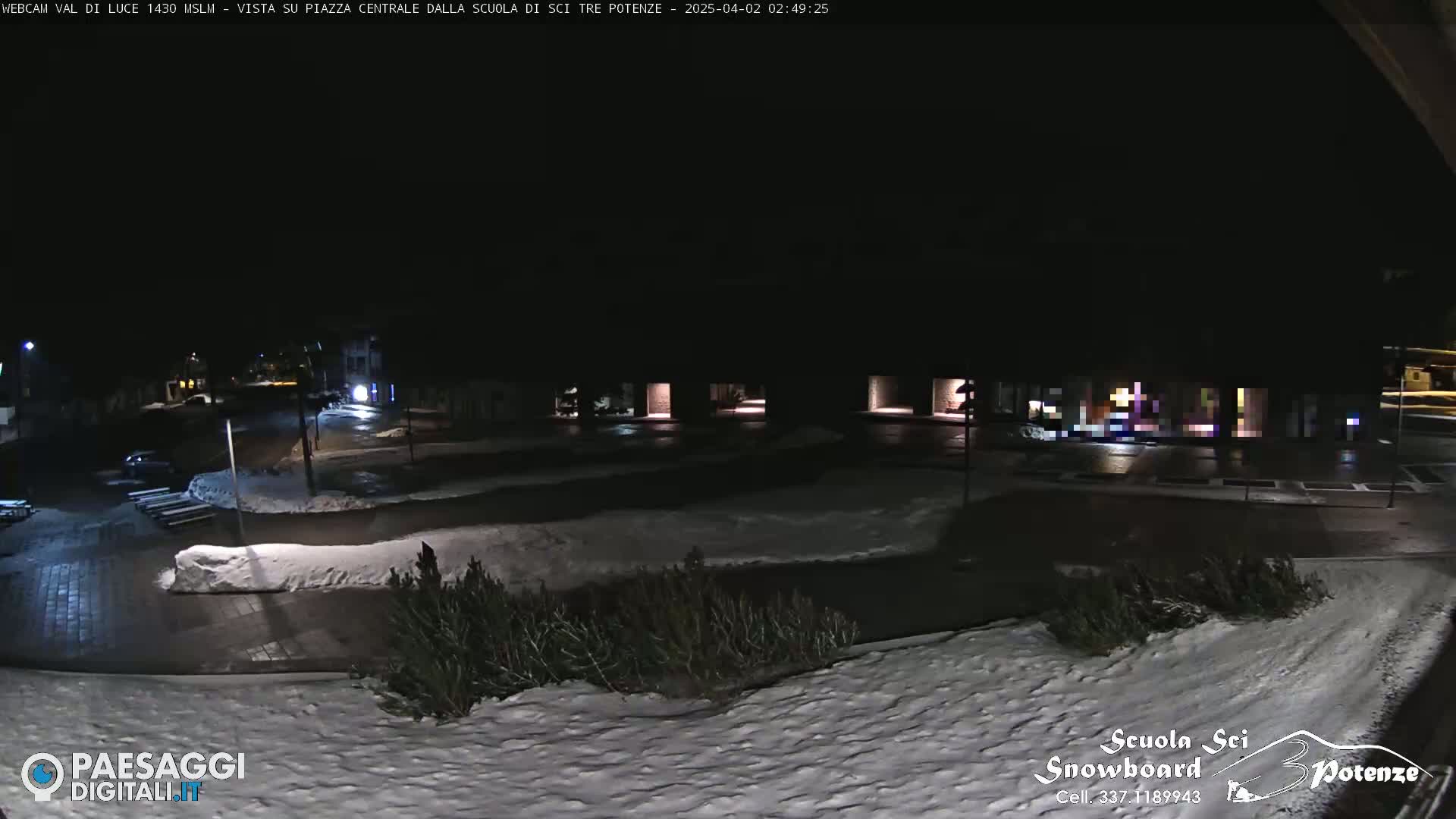 A nighttime view of a snow-covered plaza and parking lot, illuminated by streetlights, with buildings in the background.