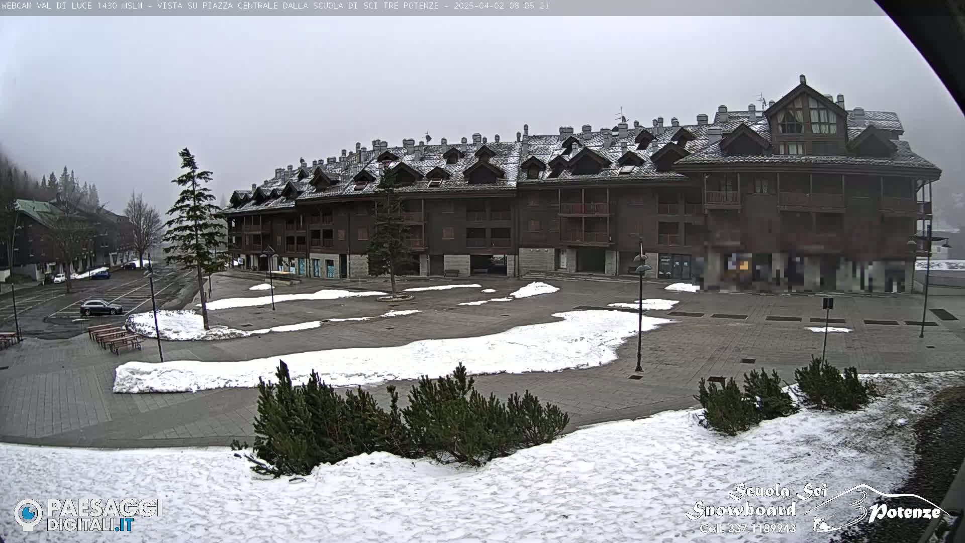 A snow-dusted town square sits before a large, multi-story brown building under an overcast sky.