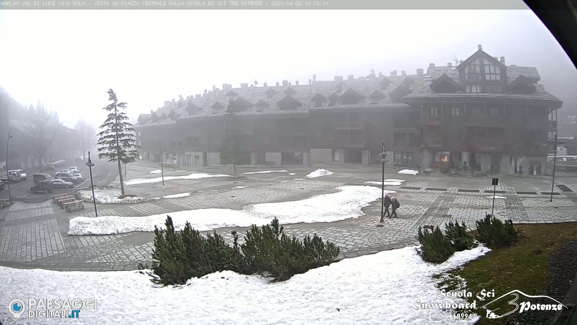 A foggy, snow-dusted plaza in front of a large multi-story building, with a few cars parked and people walking.