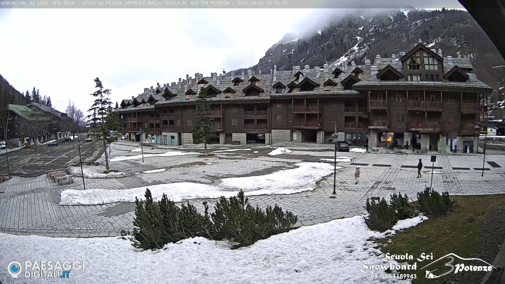 A stone plaza with patches of snow surrounds a multi-story brown building, set against a backdrop of mountains under an overcast sky.