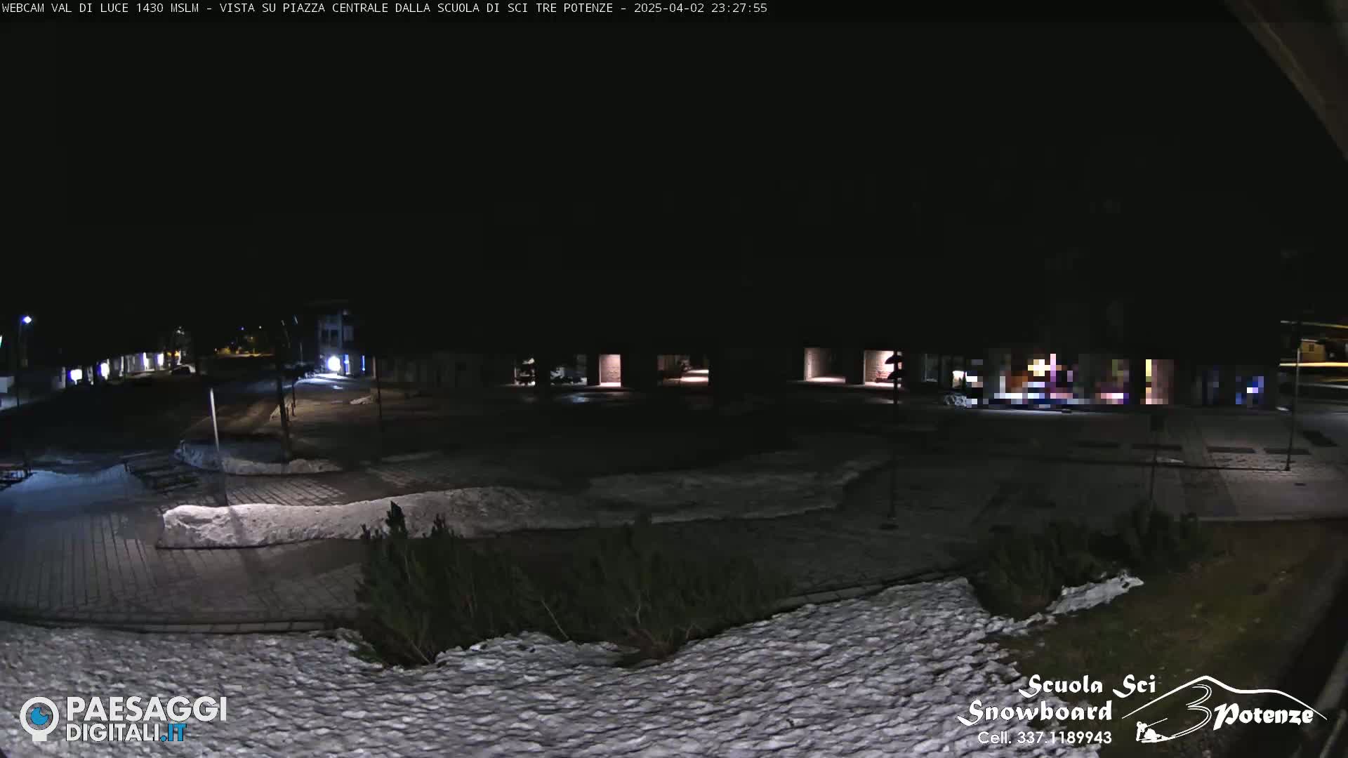 A nighttime view of a snow-dusted plaza and street, illuminated by sparse lights.
