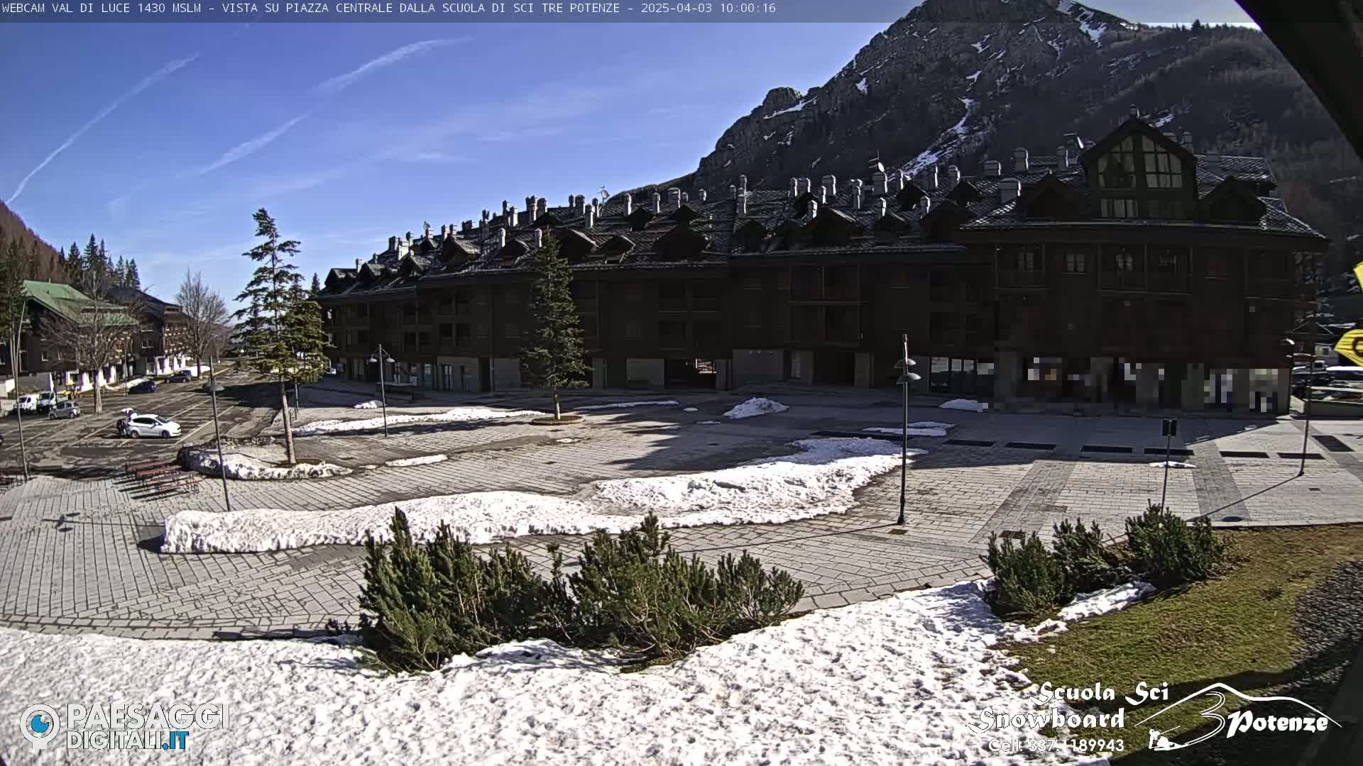 A sunny, snow-dusted plaza sits before a large, multi-story building nestled against a mountain.