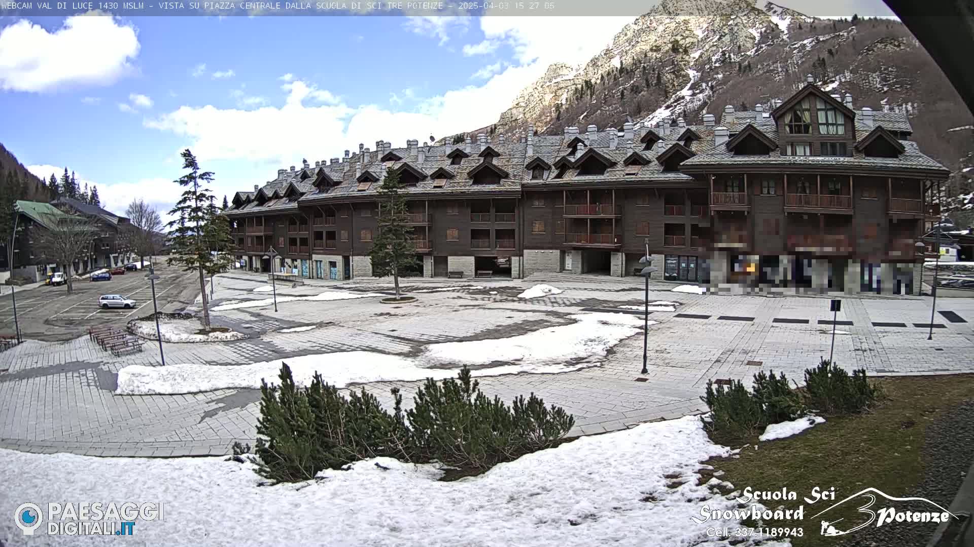 A partly snow-covered plaza sits in front of a large, multi-story brown building nestled in a mountain valley under a mostly sunny sky.