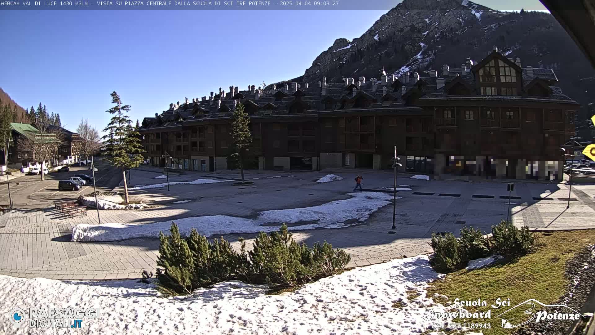 A mostly sunny, snow-dusted plaza is situated in front of a multi-story building, backed by mountains.