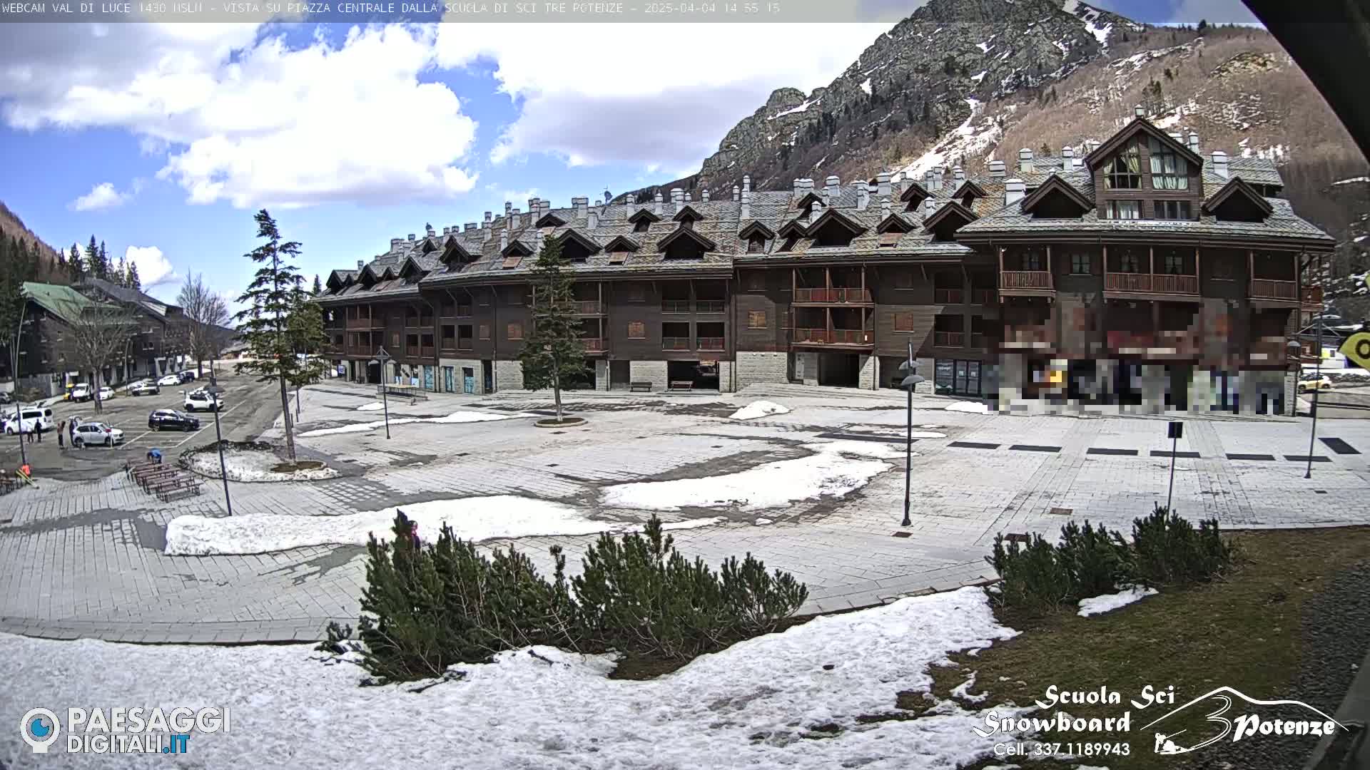 A large, multi-story building in a mountain town square partially covered in snow under a partly cloudy sky.