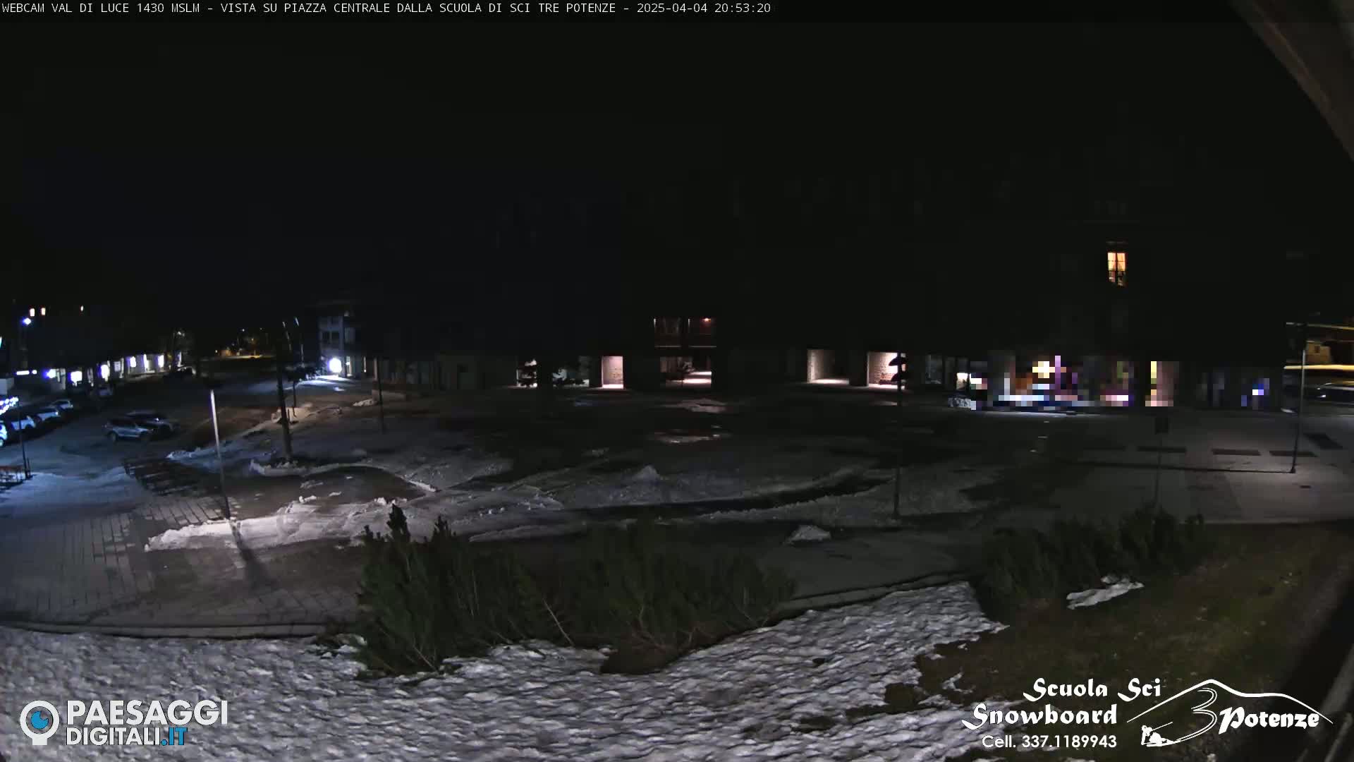A nighttime view of a snow-dusted town square, with patches of snow and ice visible on the ground and parked cars in the distance.