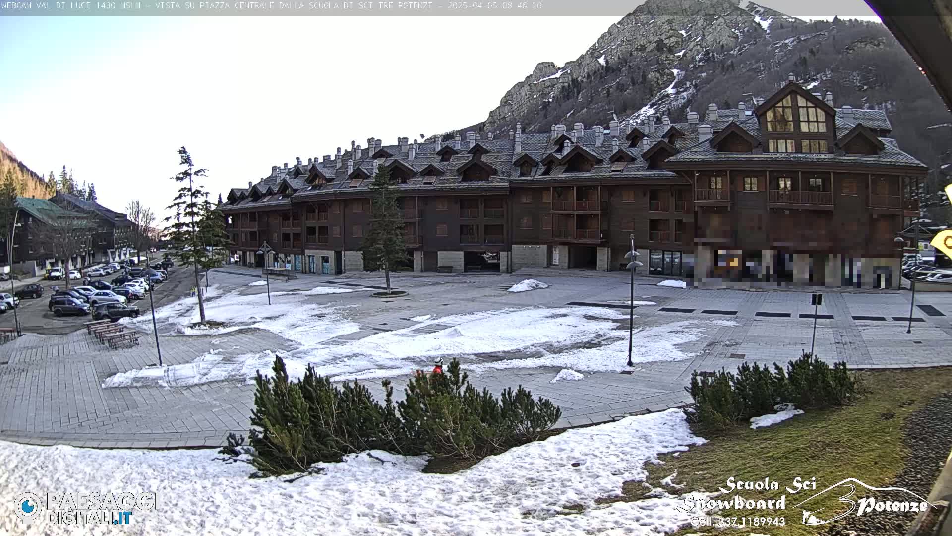 A snow-dusted plaza sits in front of a large, multi-story brown building nestled in a mountain valley under a mostly clear sky.