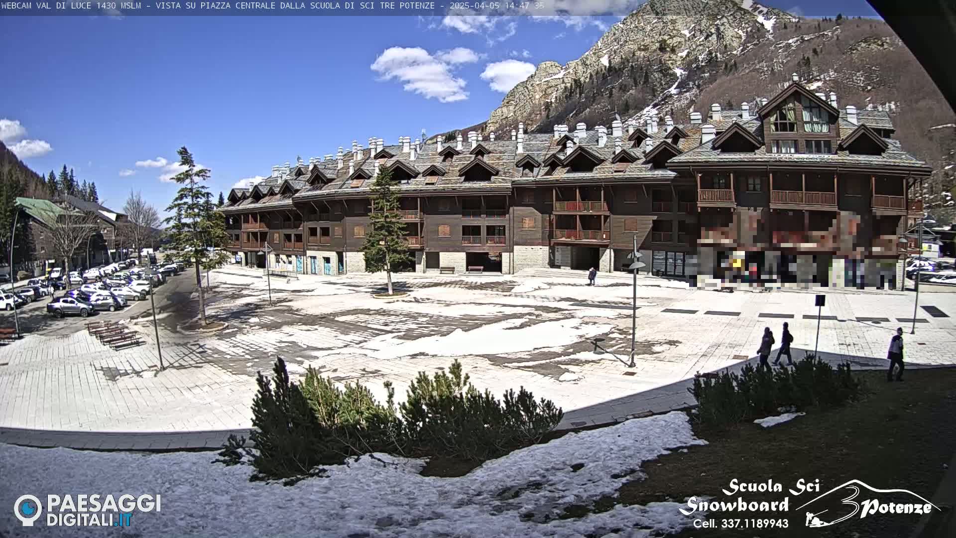 A mostly sunny day shows a large, paved town square with patches of snow, partially surrounded by a multi-story brown building against a backdrop of mountains.