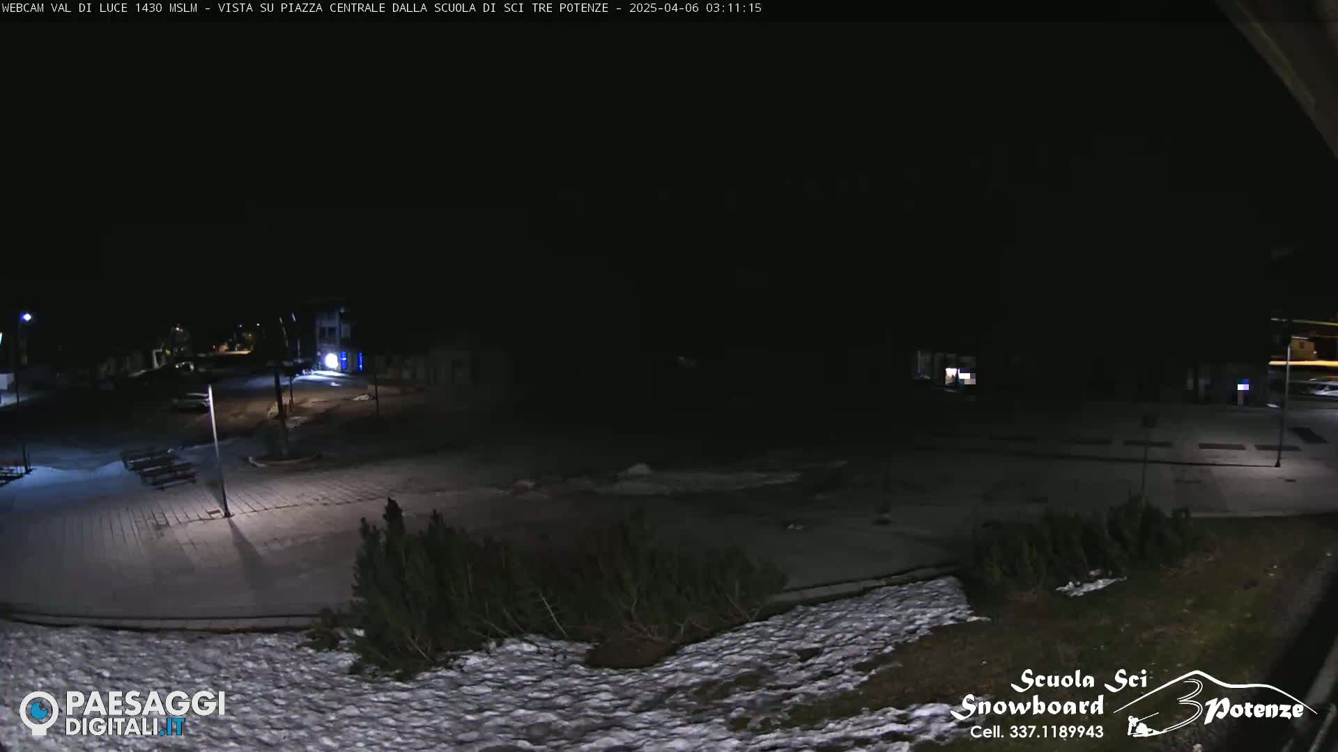 A nighttime view of a snow-dusted plaza, sparsely lit, with some small shrubs and buildings visible in the distance.