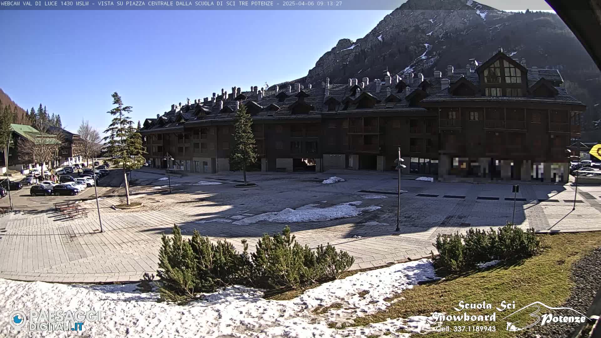 A sunny day in a mountain town shows a paved plaza with a few patches of snow, surrounded by a multi-story brown building and parked cars.