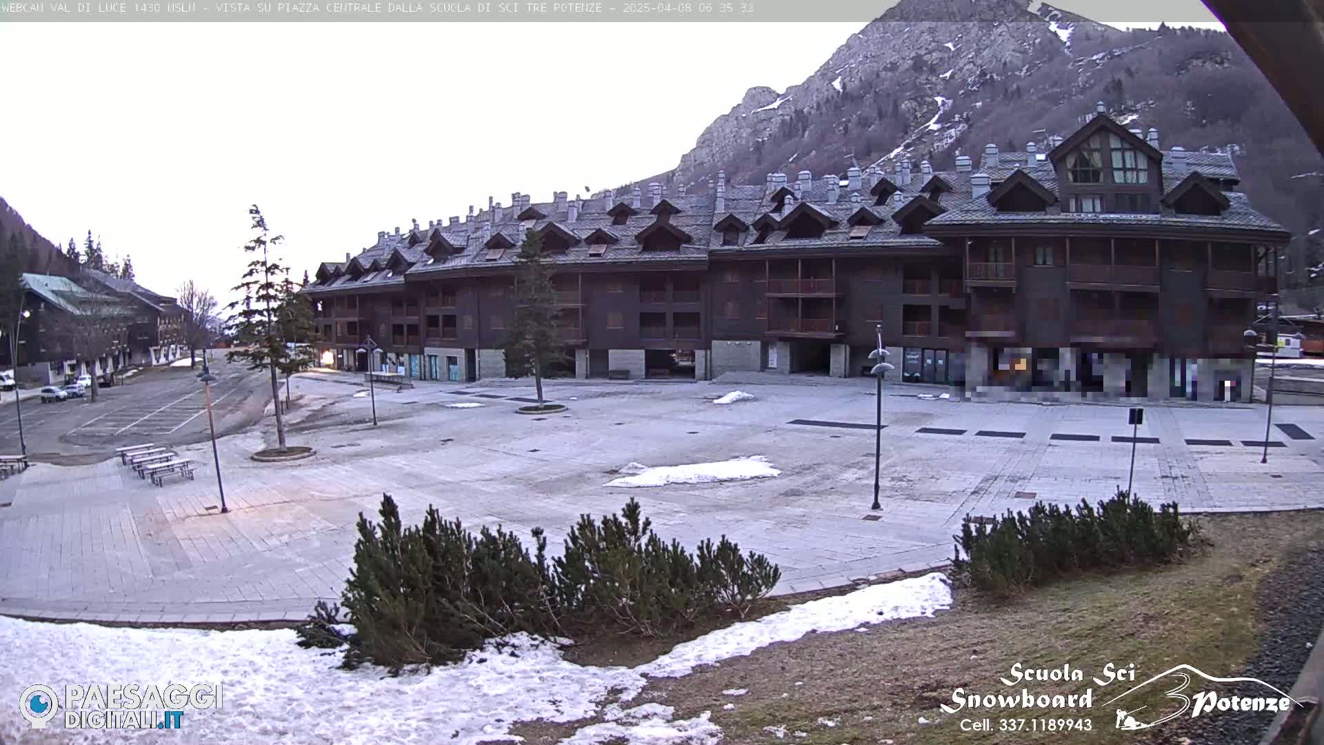 A large, empty plaza in front of a multi-story brown building is lightly dusted with snow, with mountains in the background under a cloudy sky.