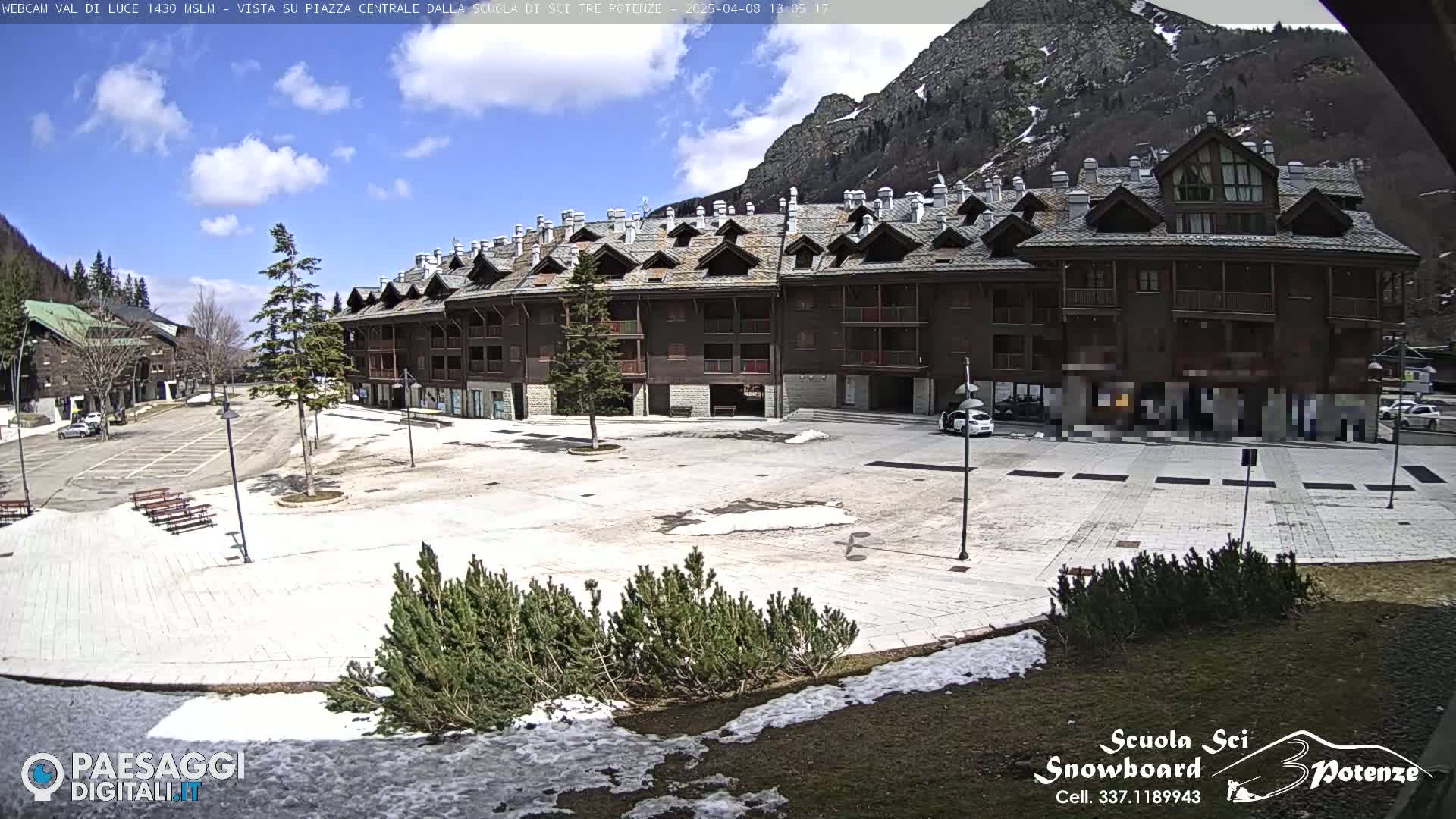 A mostly clear, sunny day shows a large, mostly empty plaza in front of a multi-story brown building, with some remaining snow patches on the ground and in the background, mountains are visible.